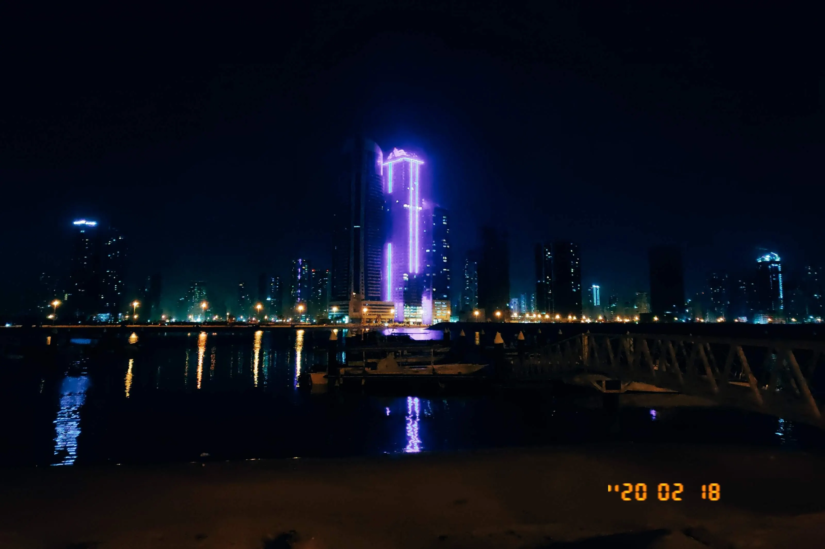 A waterfront tower at night with violet vertical lighting, the rest of the Dubai skyline glowing low behind it and a small dock in the foreground.