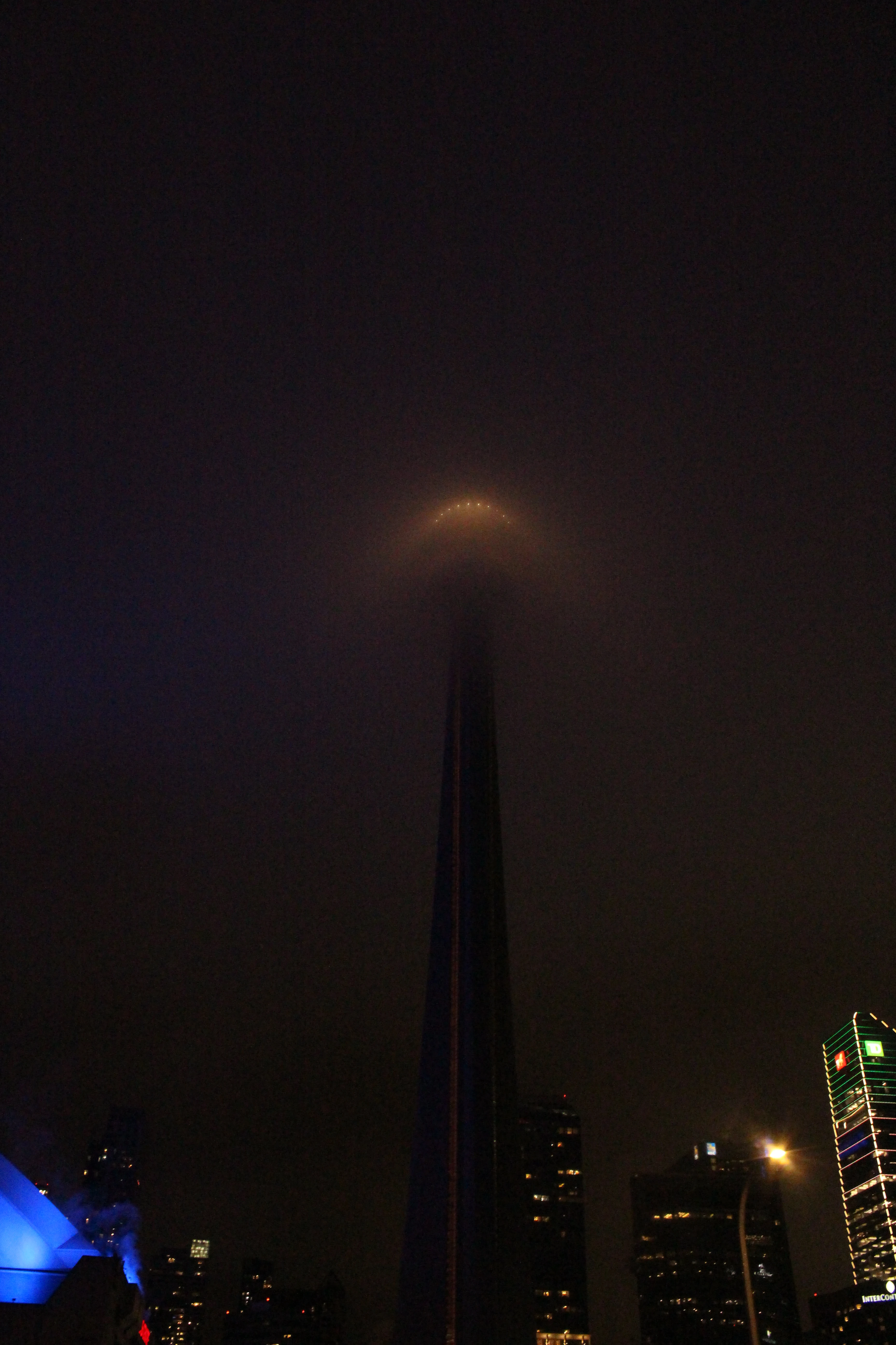CN Tower at night, half-disappeared into low fog, a soft halo around the antenna.