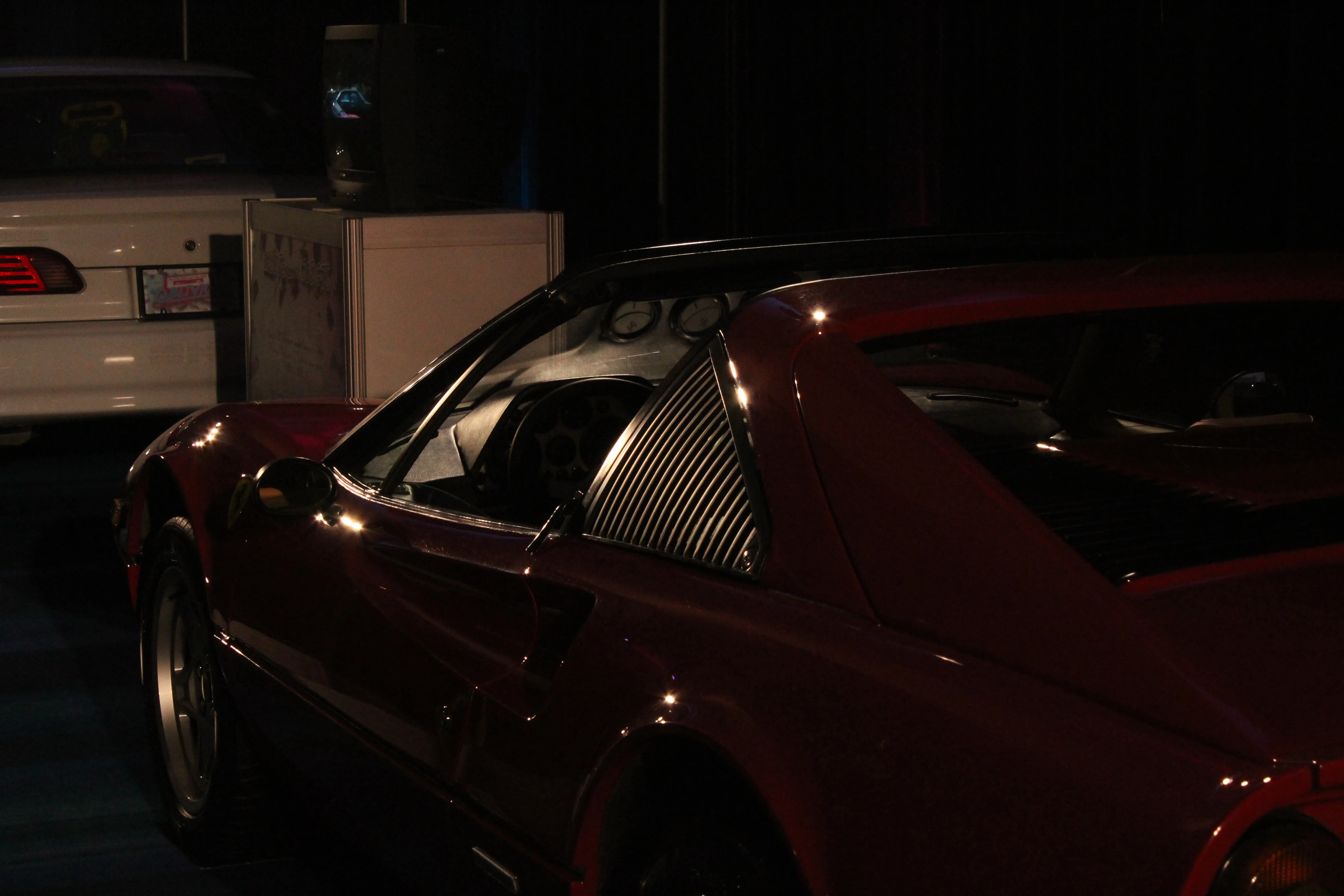 A deep red Ferrari 308 photographed from the rear three quarter under low gallery lighting, side intake grille catching a soft highlight.