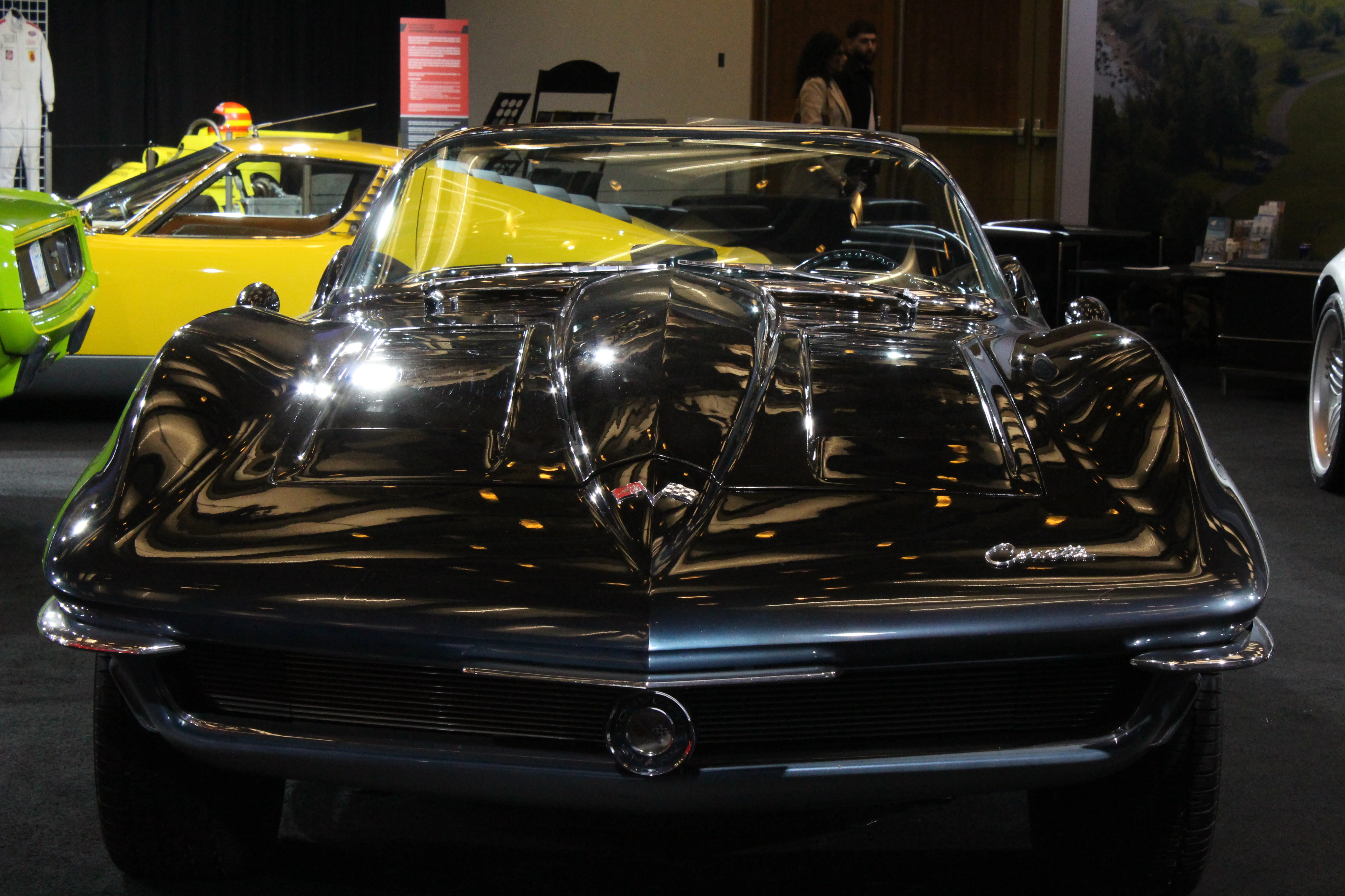 A black Chevrolet Corvette Sting Ray photographed head-on, split rear window glinting, a yellow show car visible behind on the left.