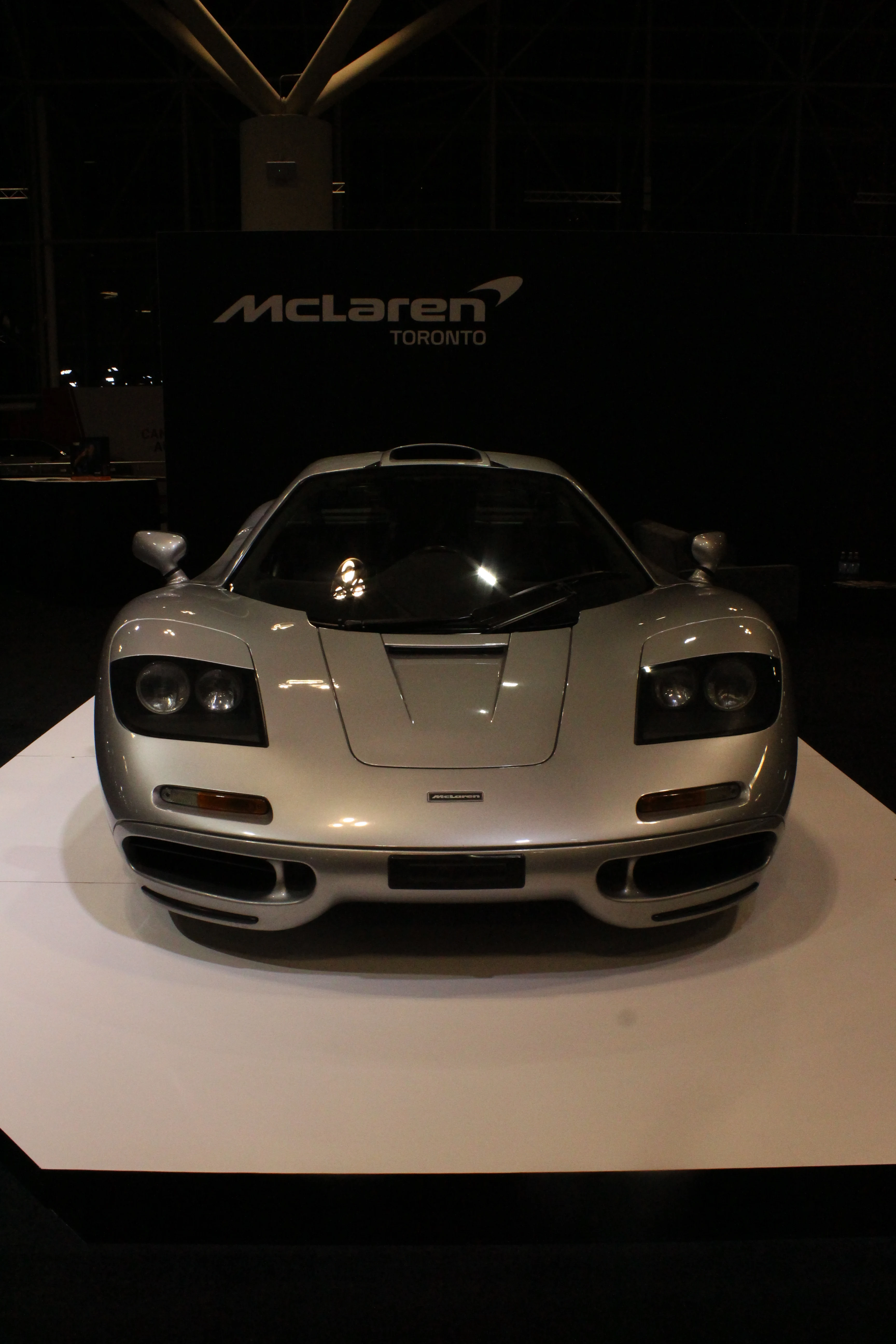 A silver McLaren coupe presented on a raised white display plinth in a darkened hall, the McLaren Toronto wordmark on the wall behind.