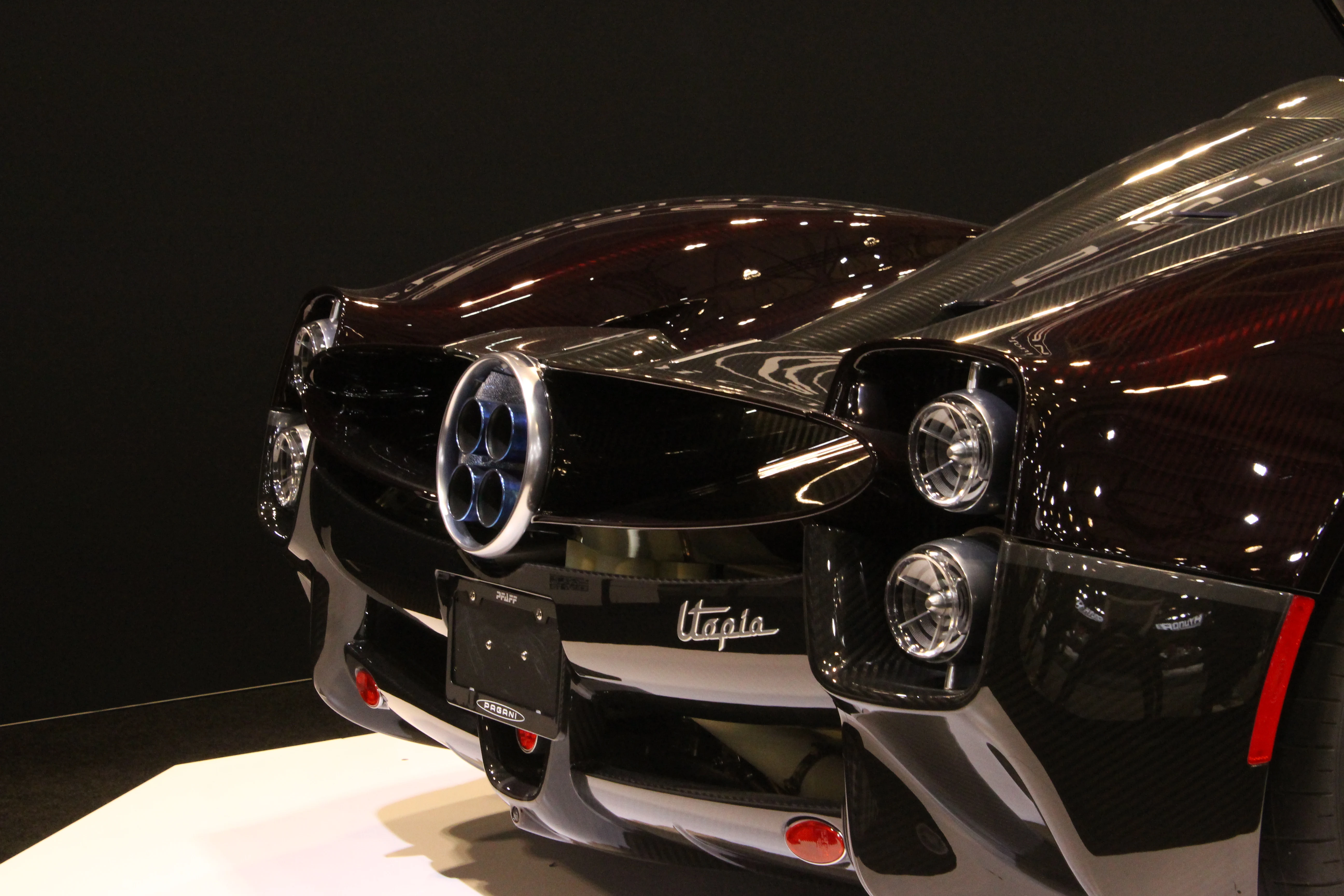 The front of a black Pagani Utopia from a low angle, exposed carbon fibre weave catching the highlights and quad headlights spotlit against shadow.