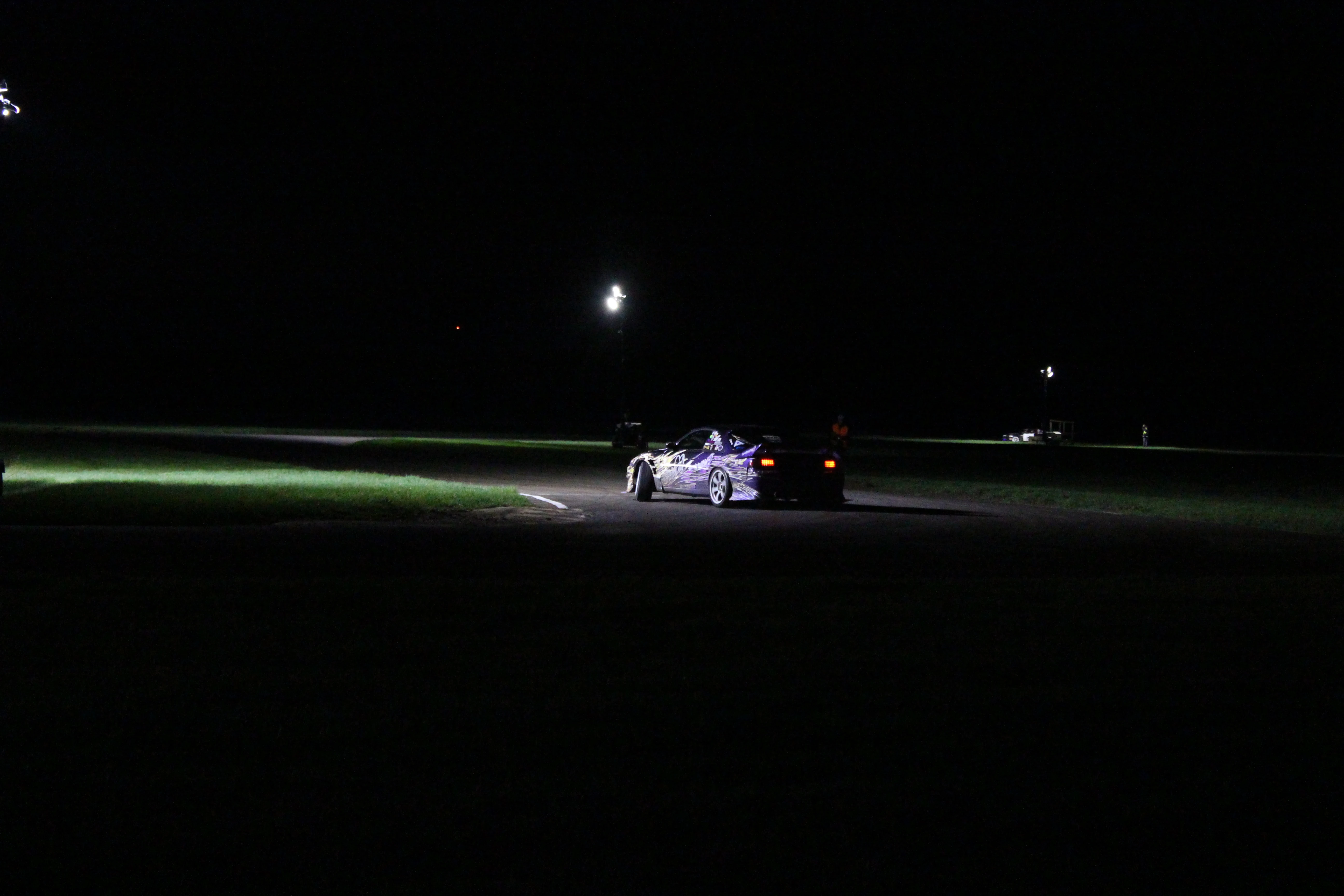 A drift car mid-corner under track floodlights at night, tyre smoke catching the light on a dark circuit.