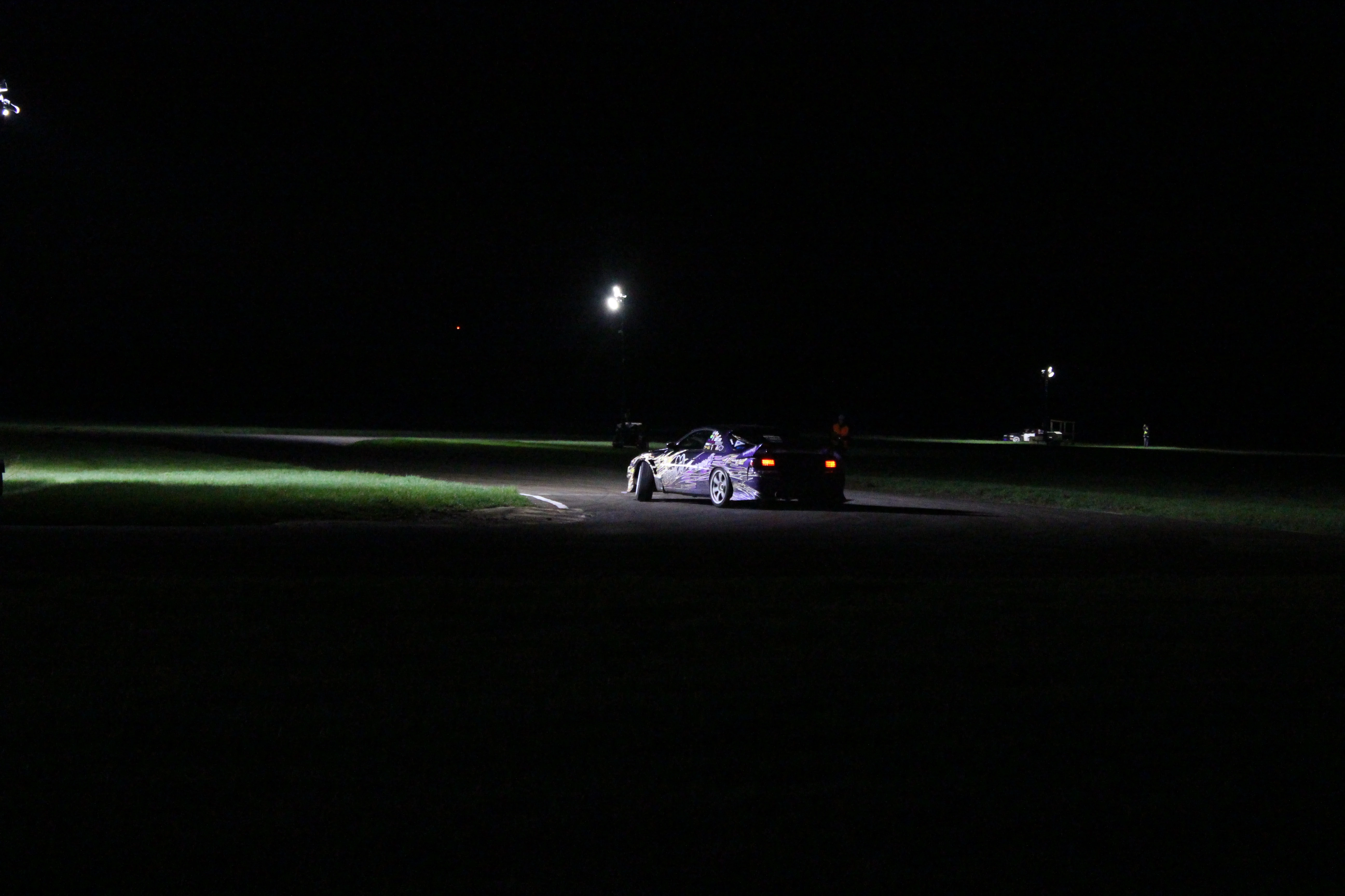A drift car mid-corner under track floodlights at night, tyre smoke catching the light on a dark circuit.