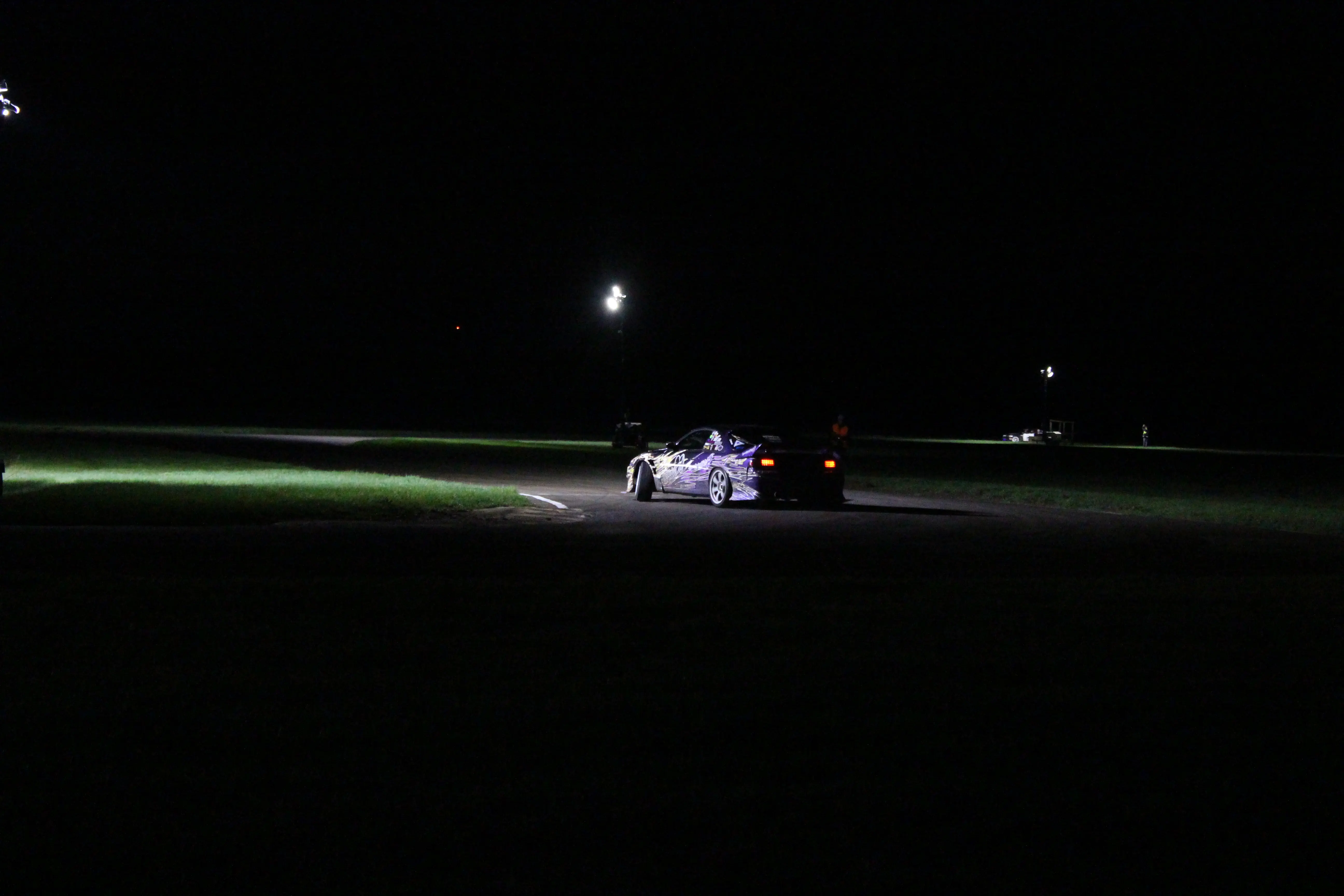 A drift car mid-corner under track floodlights at night, tyre smoke catching the light on a dark circuit.