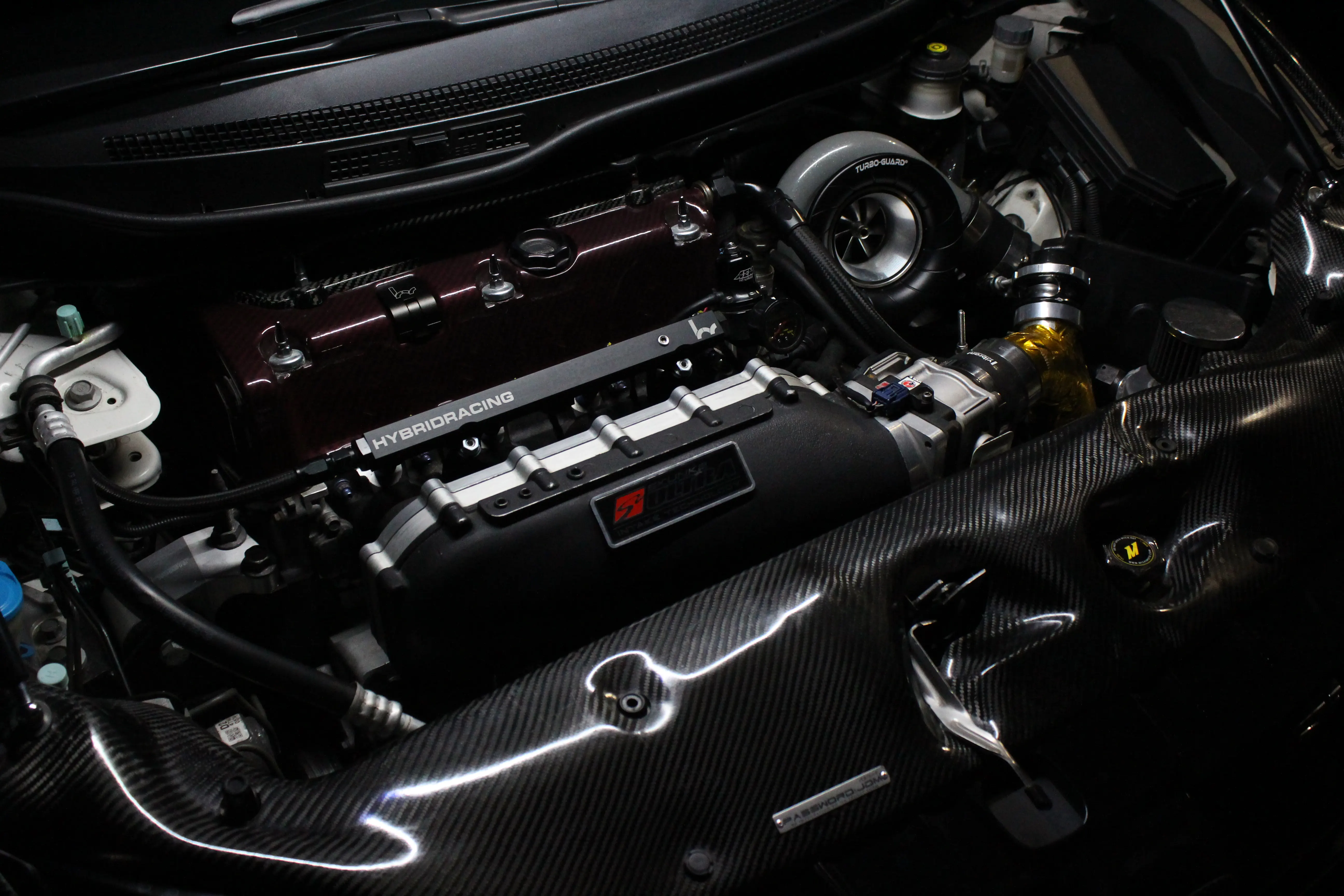A built Honda K-series engine bay with a large turbo, carbon panels, and a red valve cover under low light.
