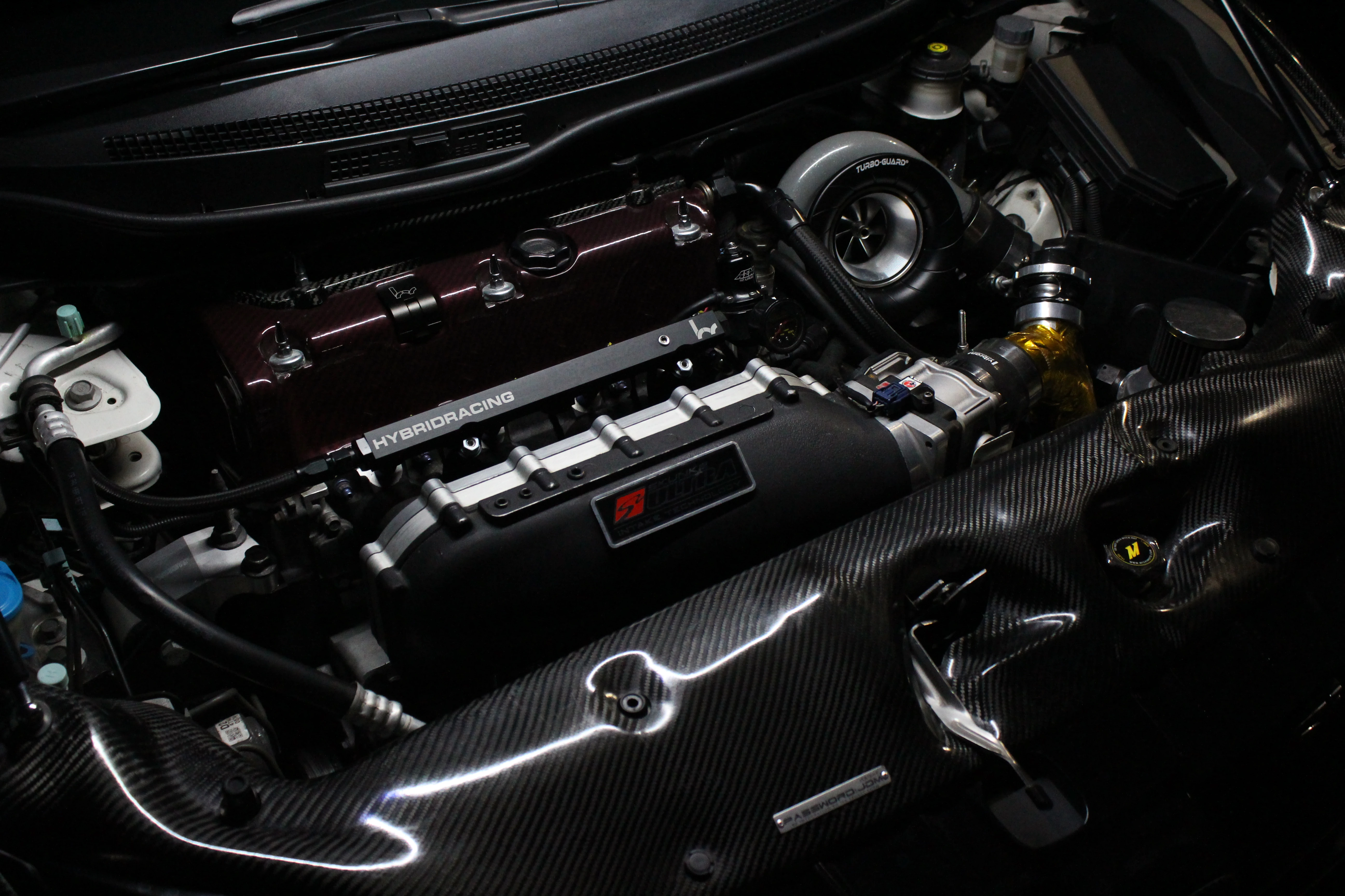 A built Honda K-series engine bay with a large turbo, carbon panels, and a red valve cover under low light.