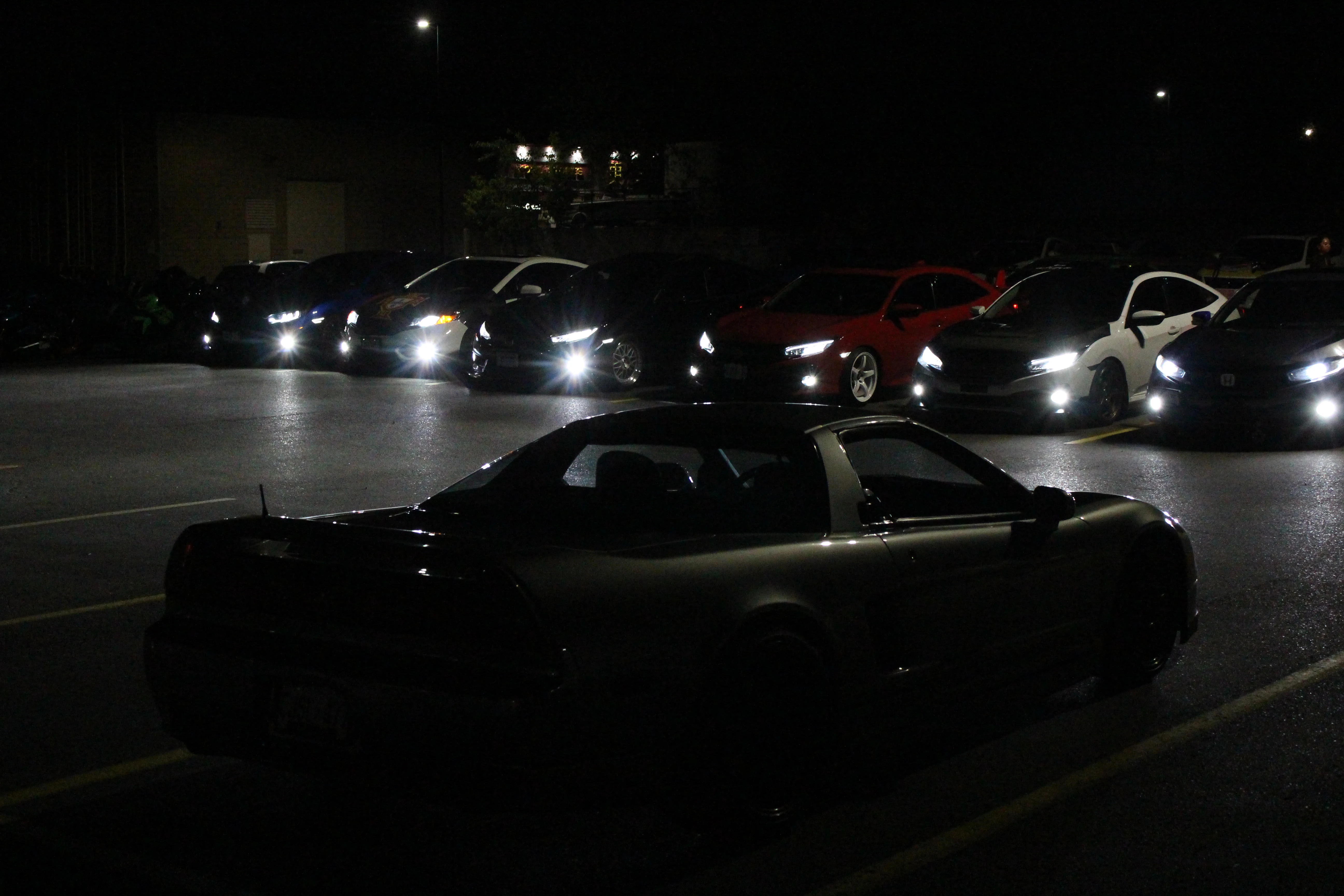 A black NSX in the foreground of a night parking-lot car meet, rows of headlights behind it.