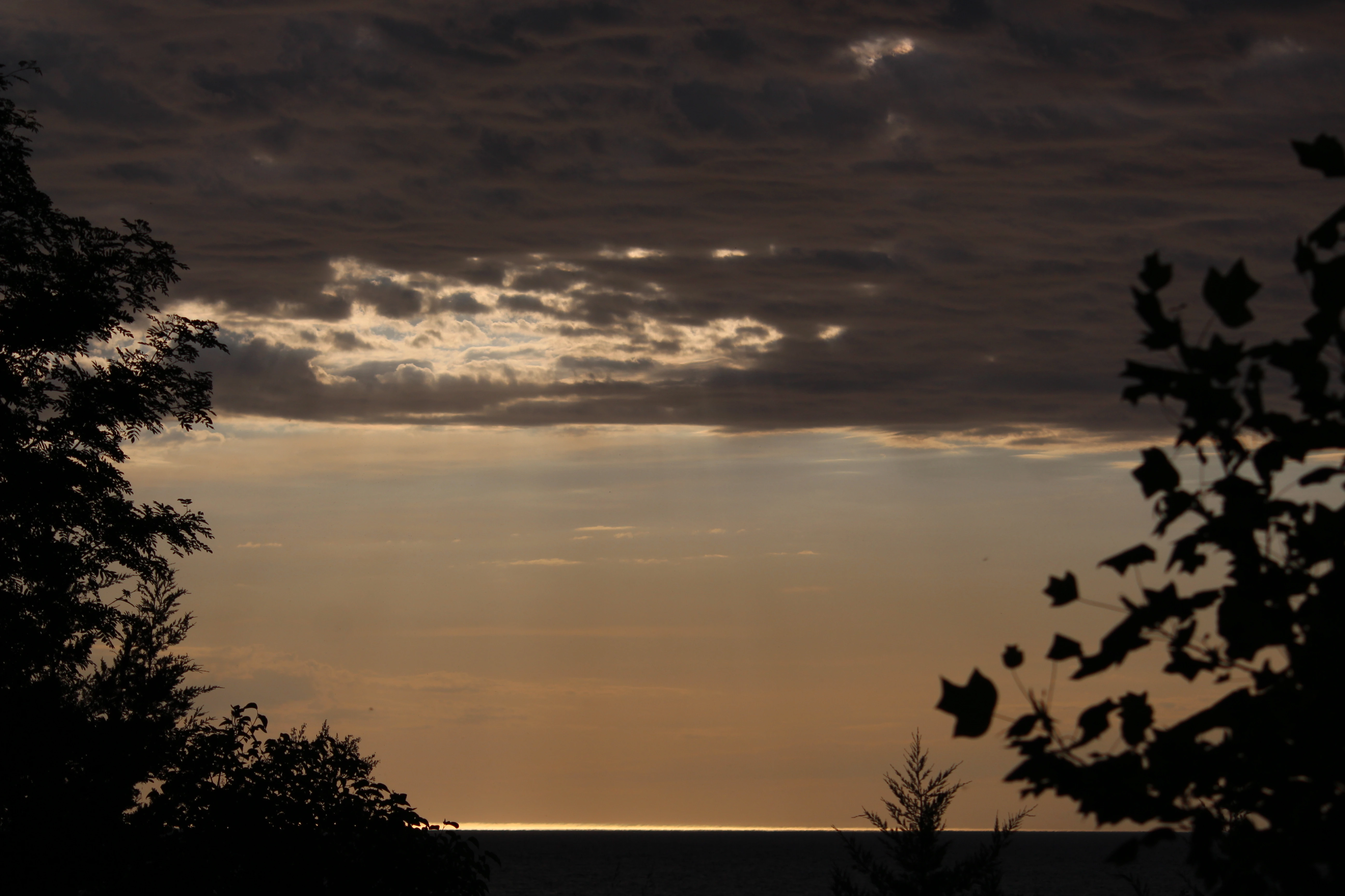 Sun breaking through heavy cloud over Lake Ontario, framed by dark silhouetted trees.