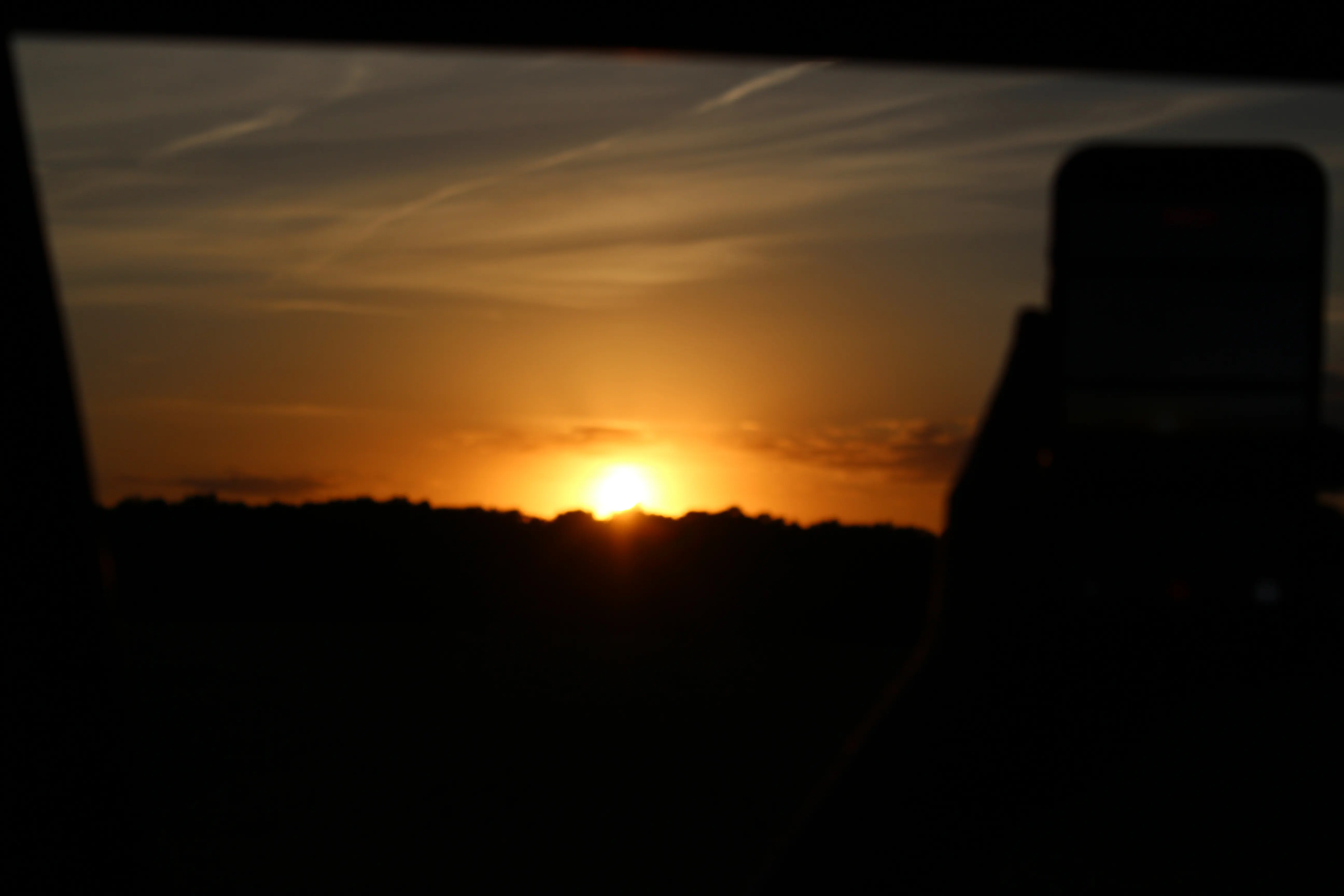 A low sun setting over a treeline, shot through a car window with a phone visible in frame.