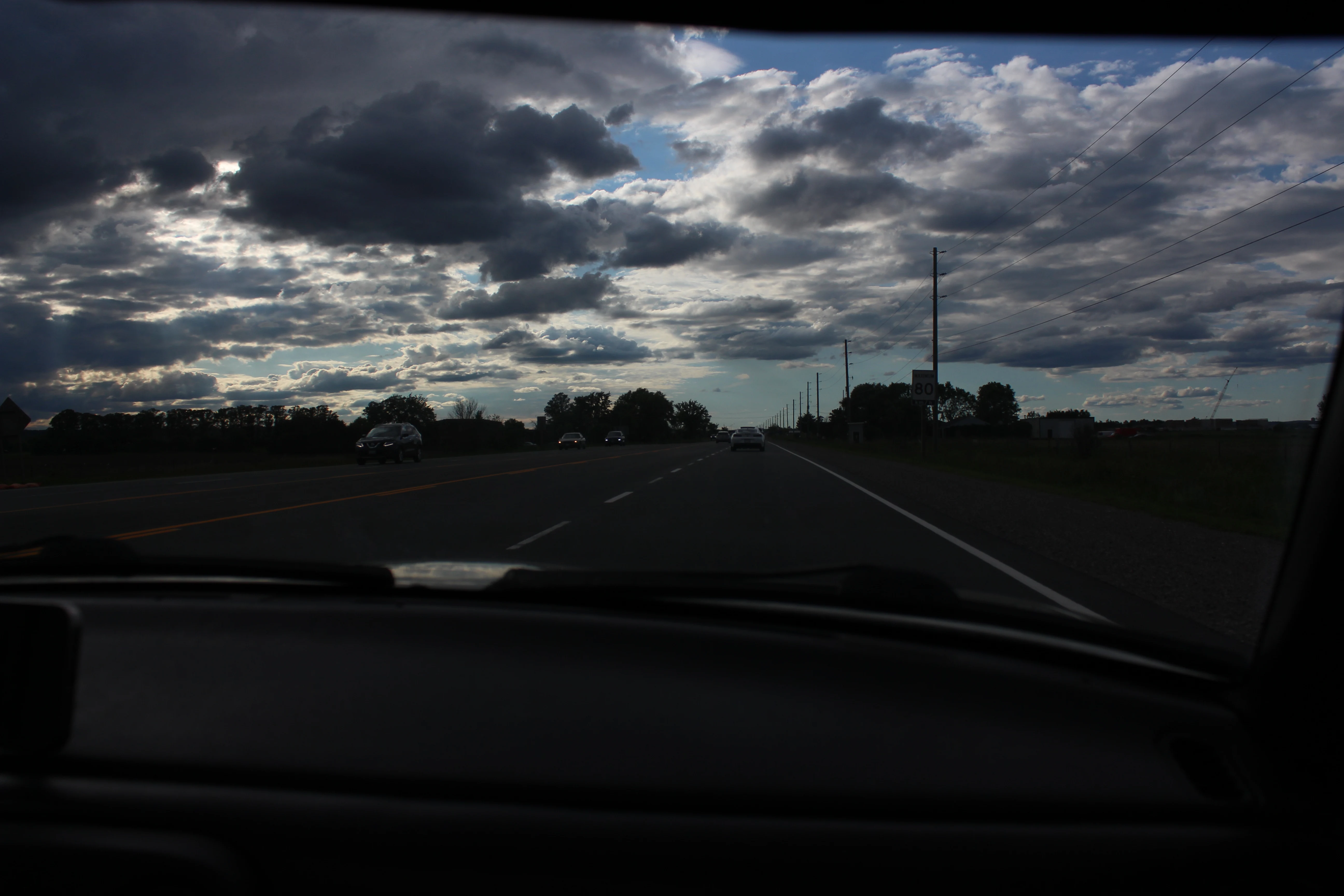 Broken clouds over an open highway seen through a car windshield, dashboard dark in the foreground.