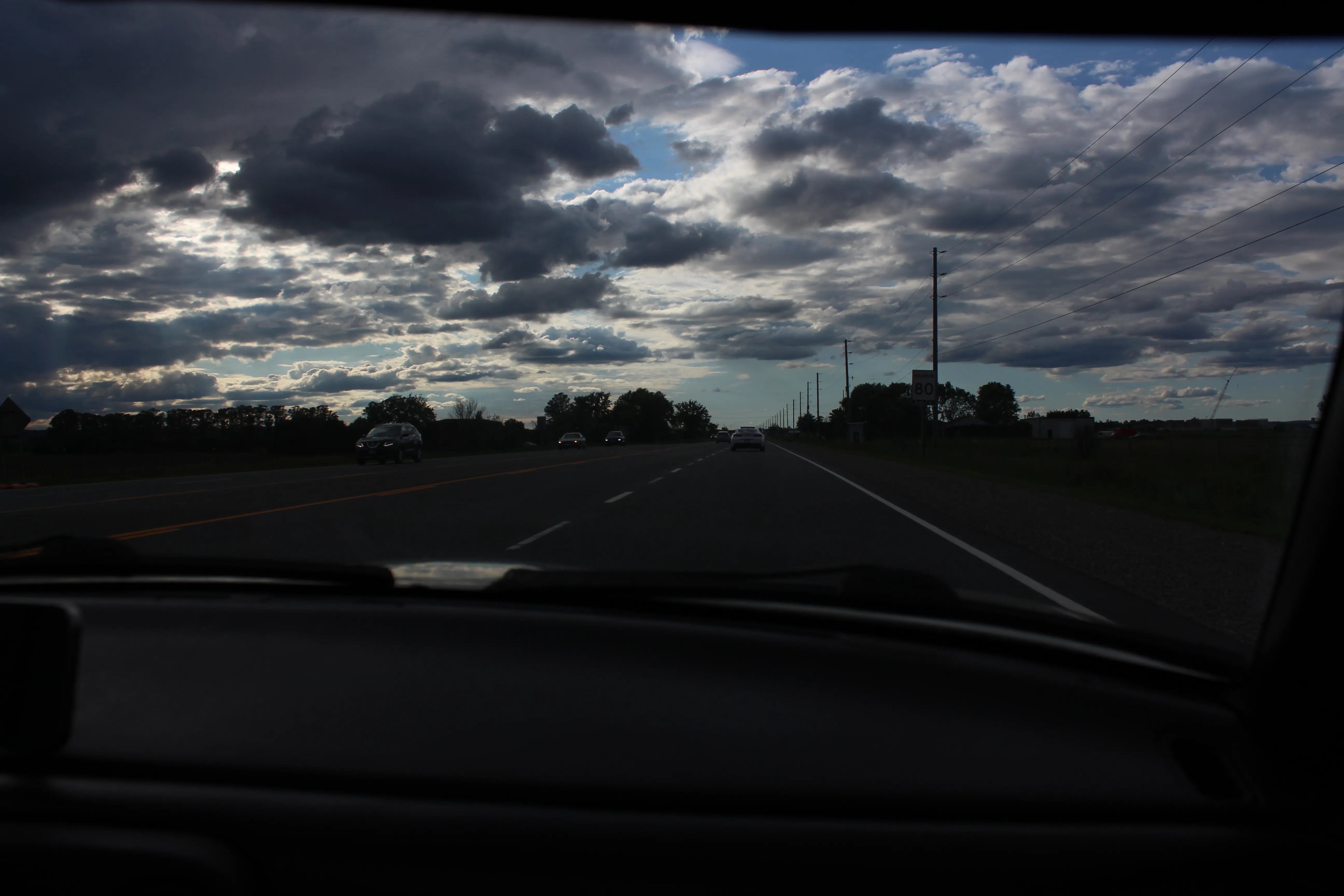 Broken clouds over an open highway seen through a car windshield, dashboard dark in the foreground.