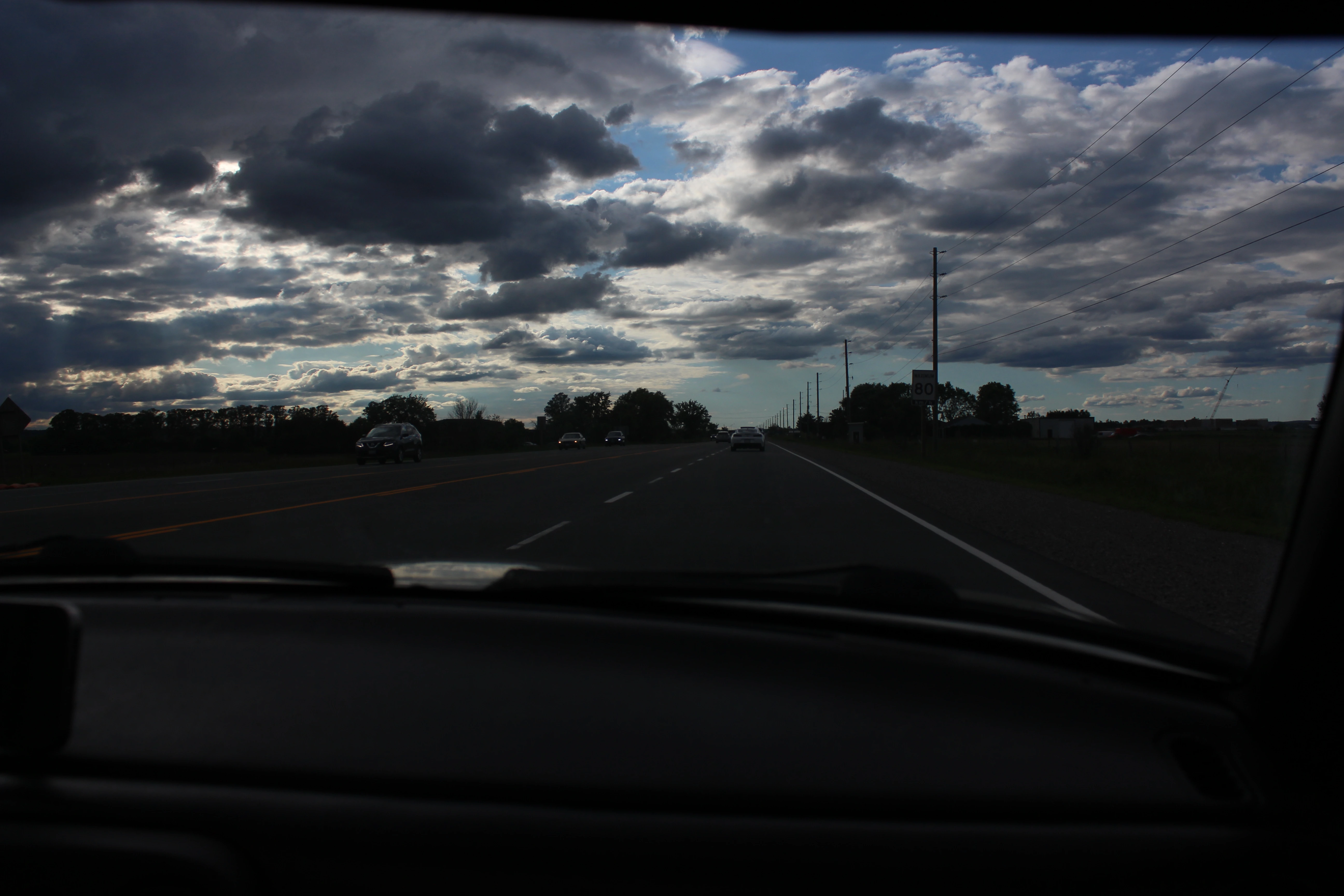 Broken clouds over an open highway seen through a car windshield, dashboard dark in the foreground.