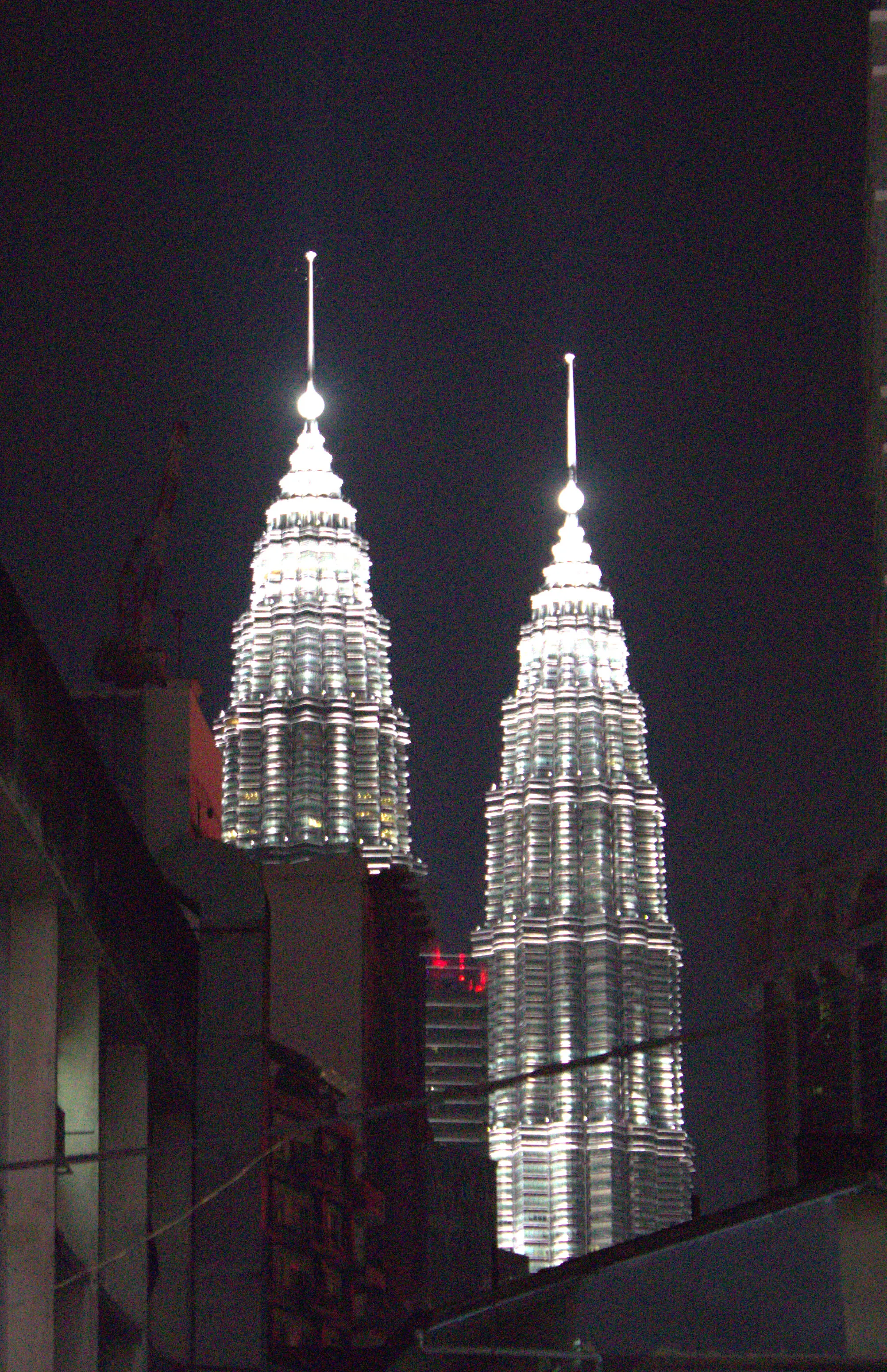 The Petronas Twin Towers at night, illuminated steel skin glowing white against a black sky, framed between darker buildings in the foreground.