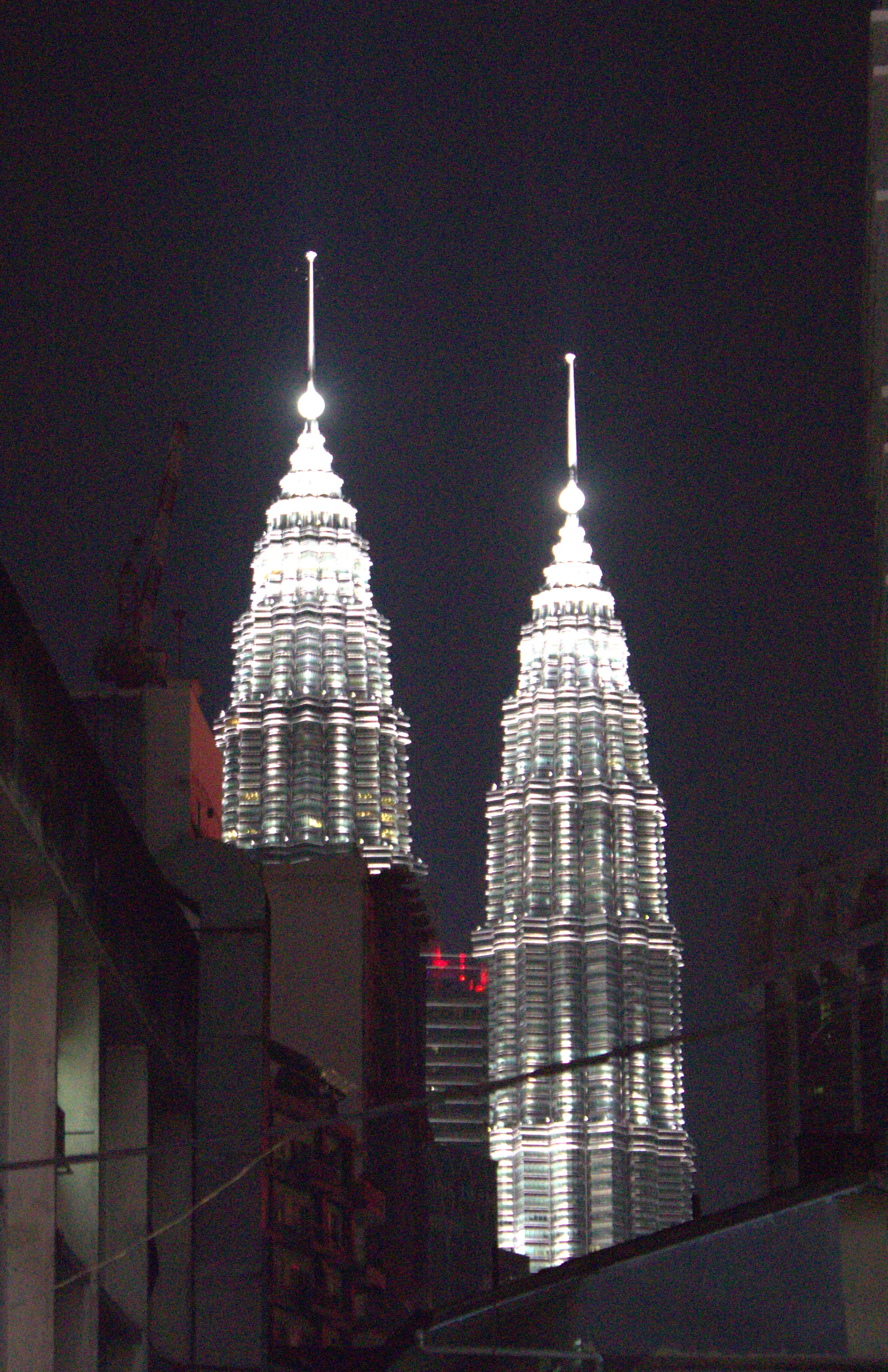 The Petronas Twin Towers at night, illuminated steel skin glowing white against a black sky, framed between darker buildings in the foreground.