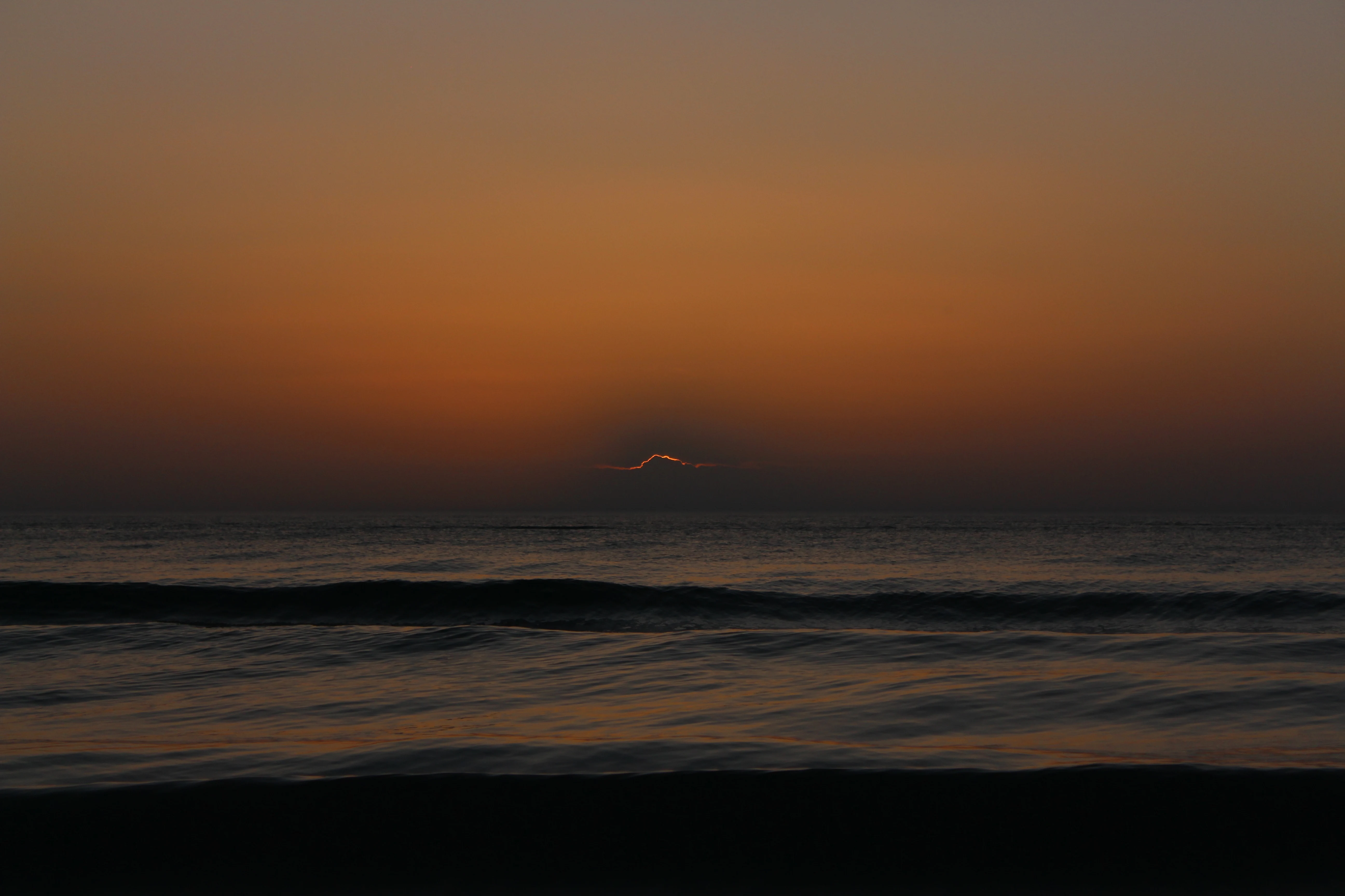 A warm amber sunset over the ocean, the sun breaking low through haze on the horizon above gentle waves.