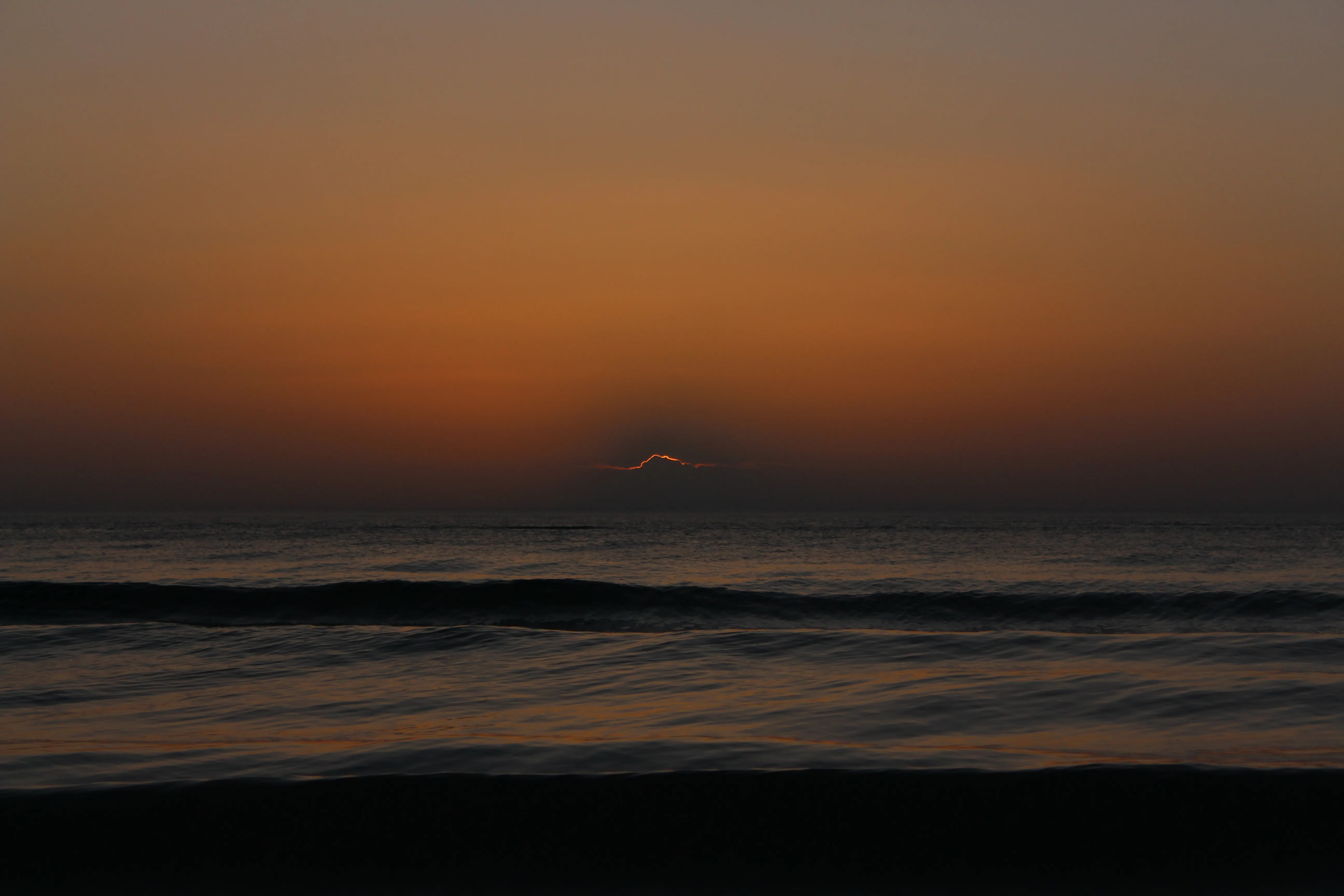A warm amber sunset over the ocean, the sun breaking low through haze on the horizon above gentle waves.