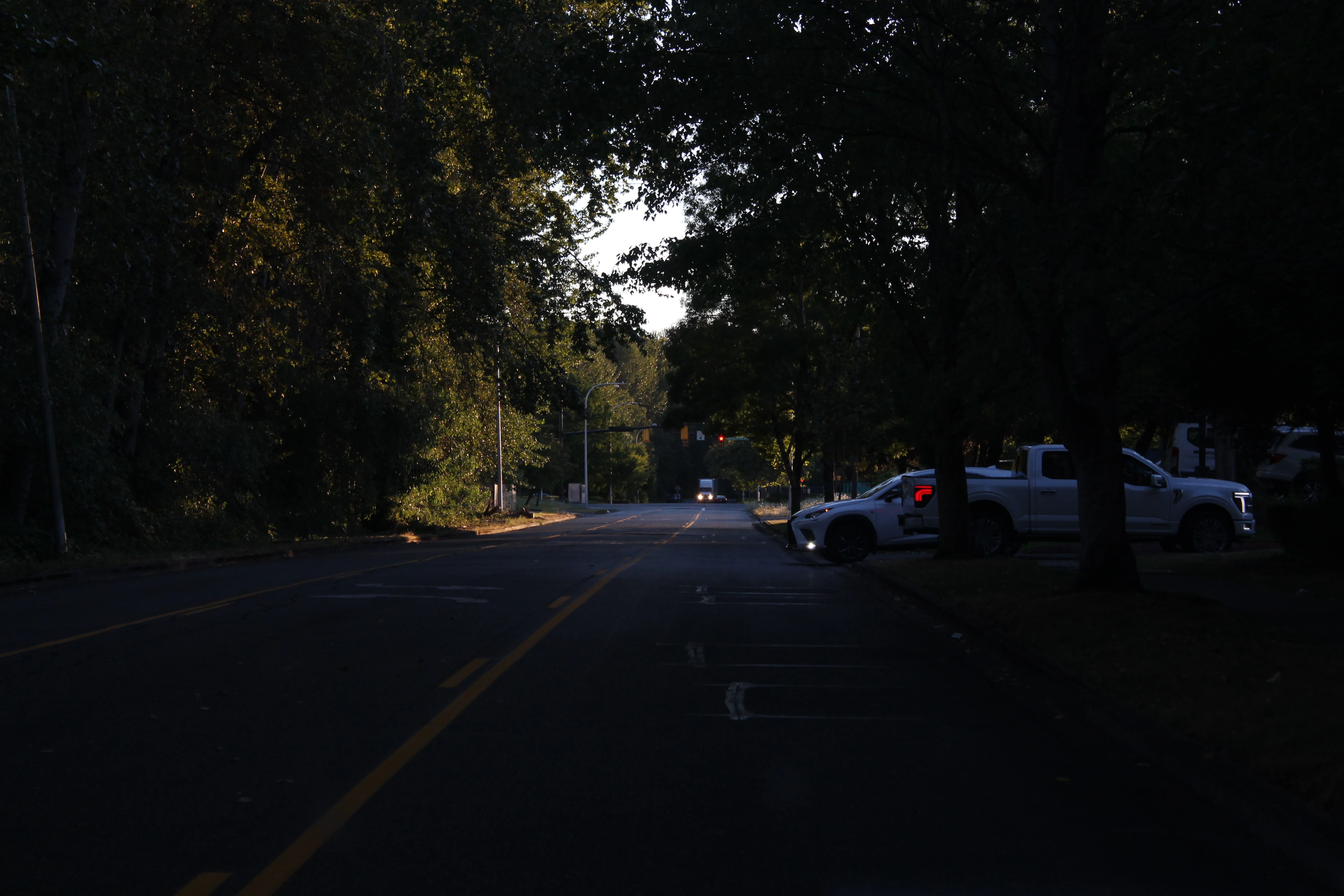 A tree-lined residential street at dusk, parked trucks along the right and the last daylight glowing through the canopy at the far end.