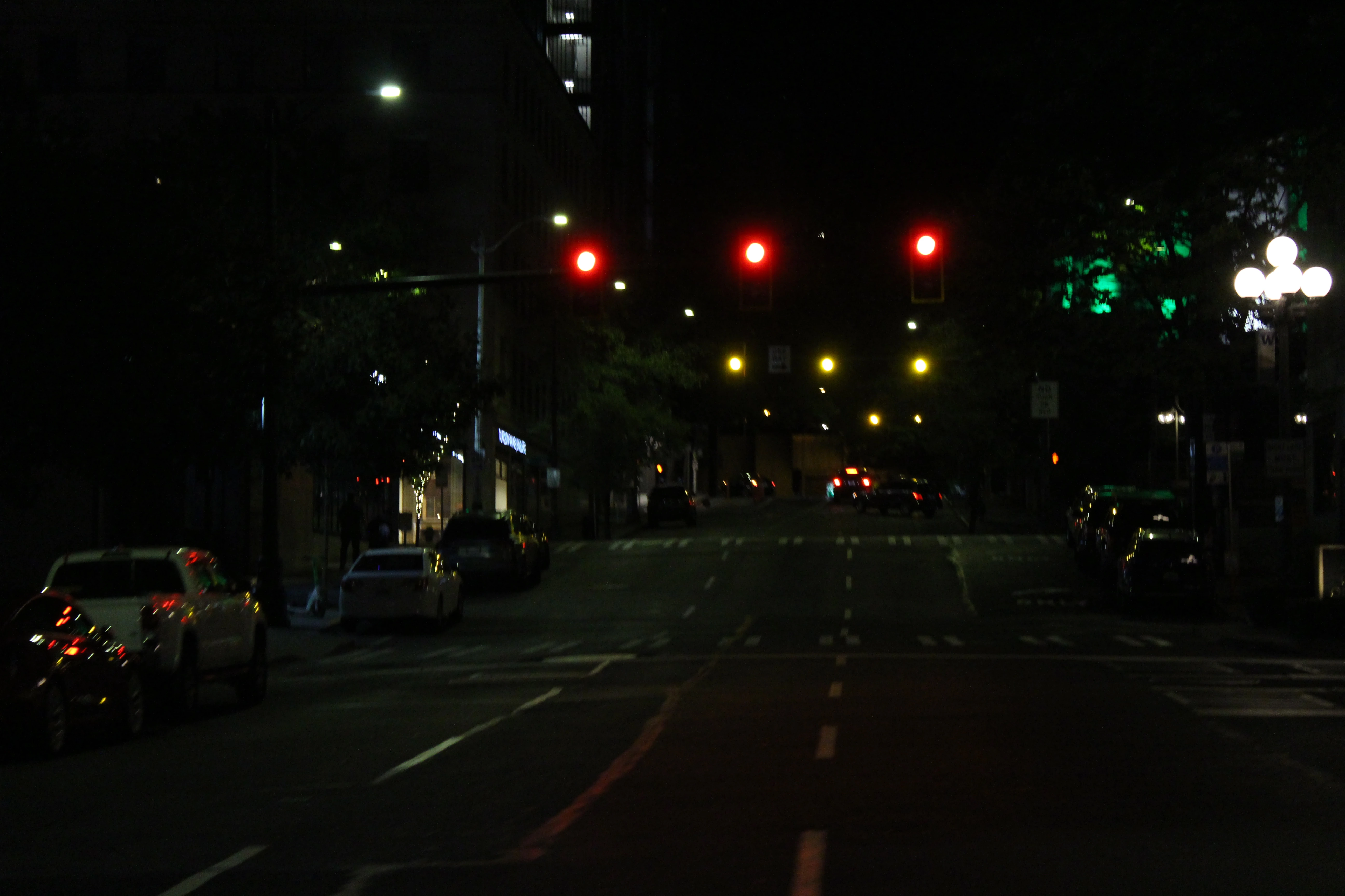 An empty downtown street at night, red traffic signals hanging over the intersection and lit globe lamps along the sidewalk.