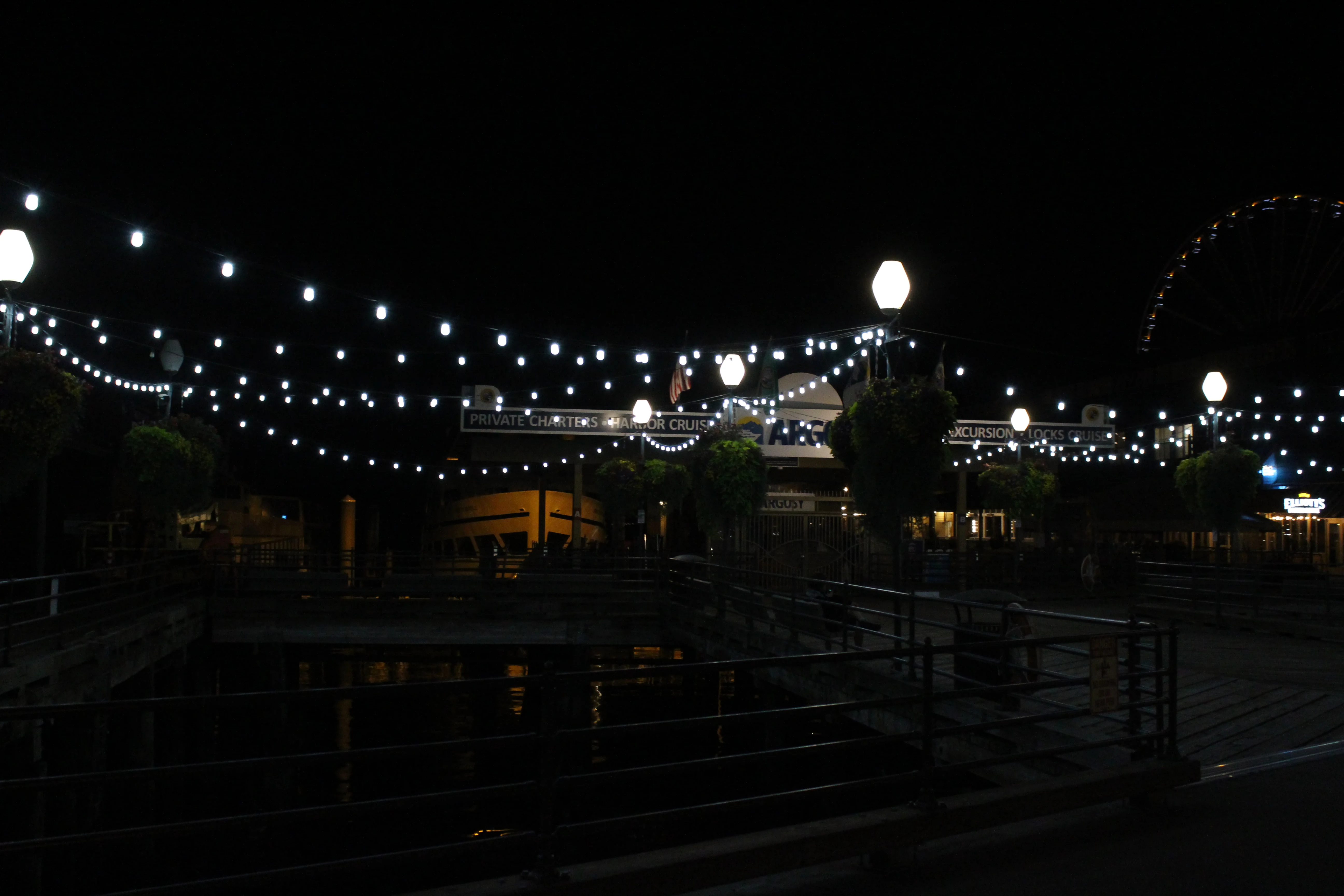 A waterfront pier strung with rows of festoon lights at night, harbor-cruise signage glowing and a Ferris wheel lit on the right.