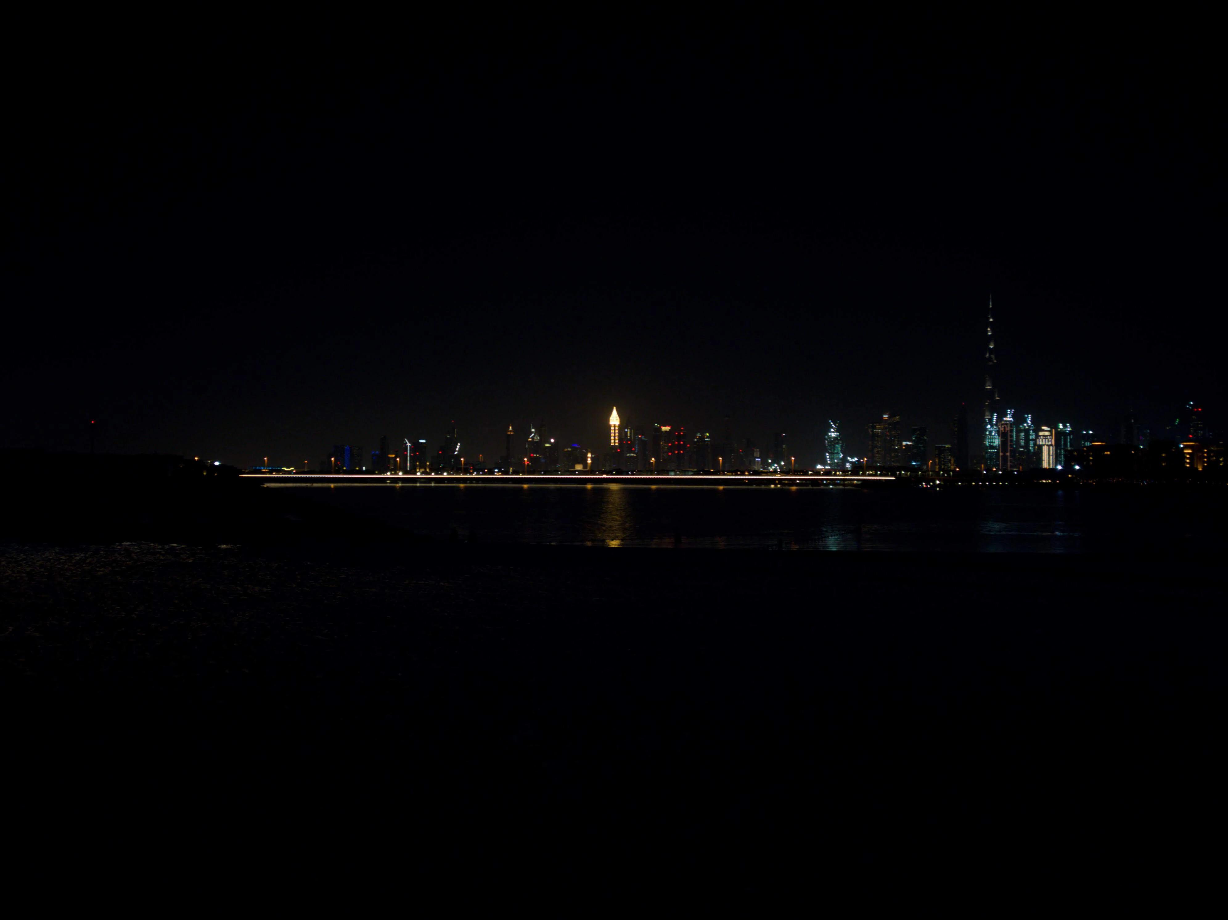 A long exposure of the Dubai skyline at night seen across dark water, the Burj Khalifa rising on the right and a streak of road lights cutting through the middle.
