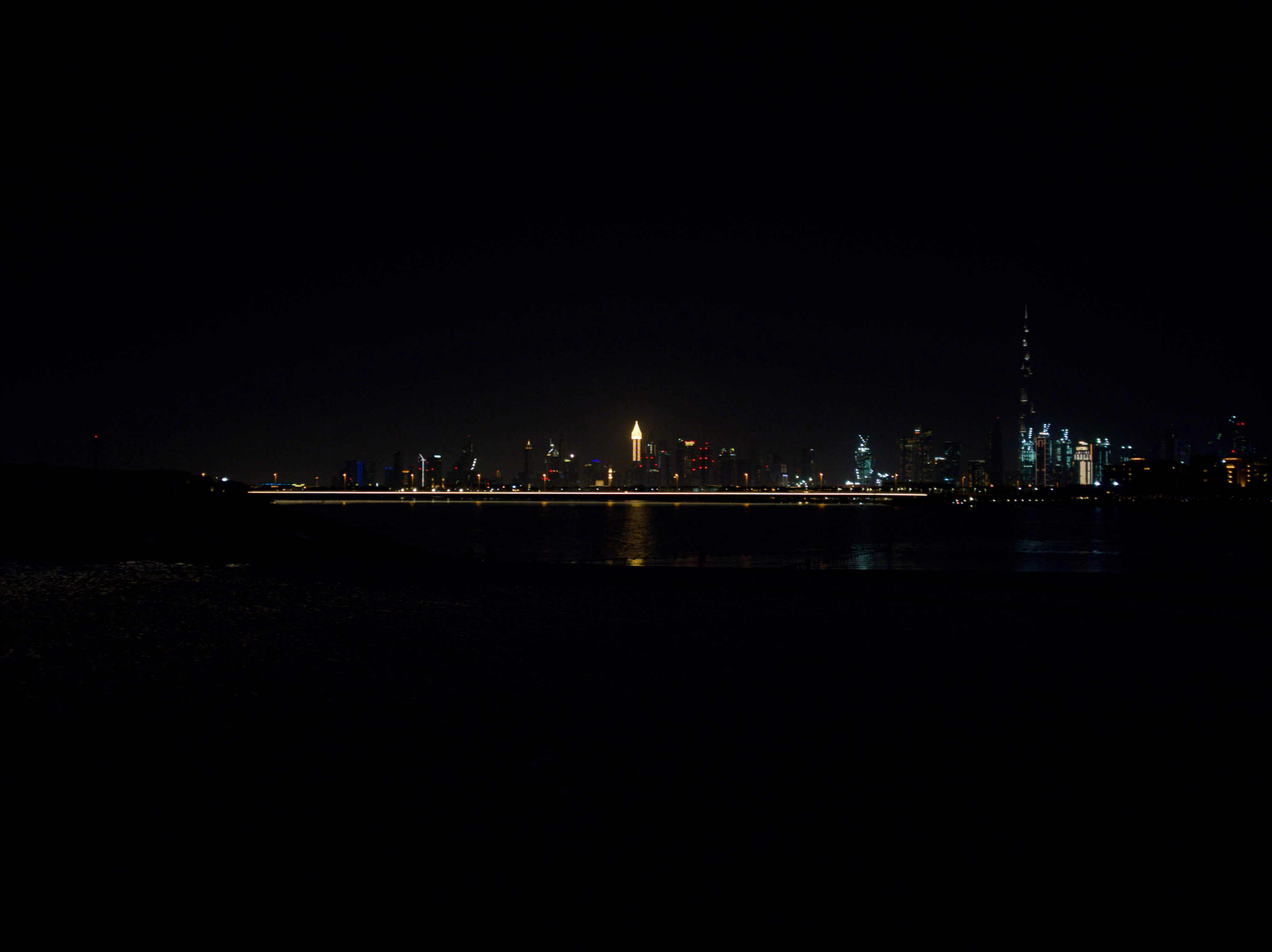 A long exposure of the Dubai skyline at night seen across dark water, the Burj Khalifa rising on the right and a streak of road lights cutting through the middle.