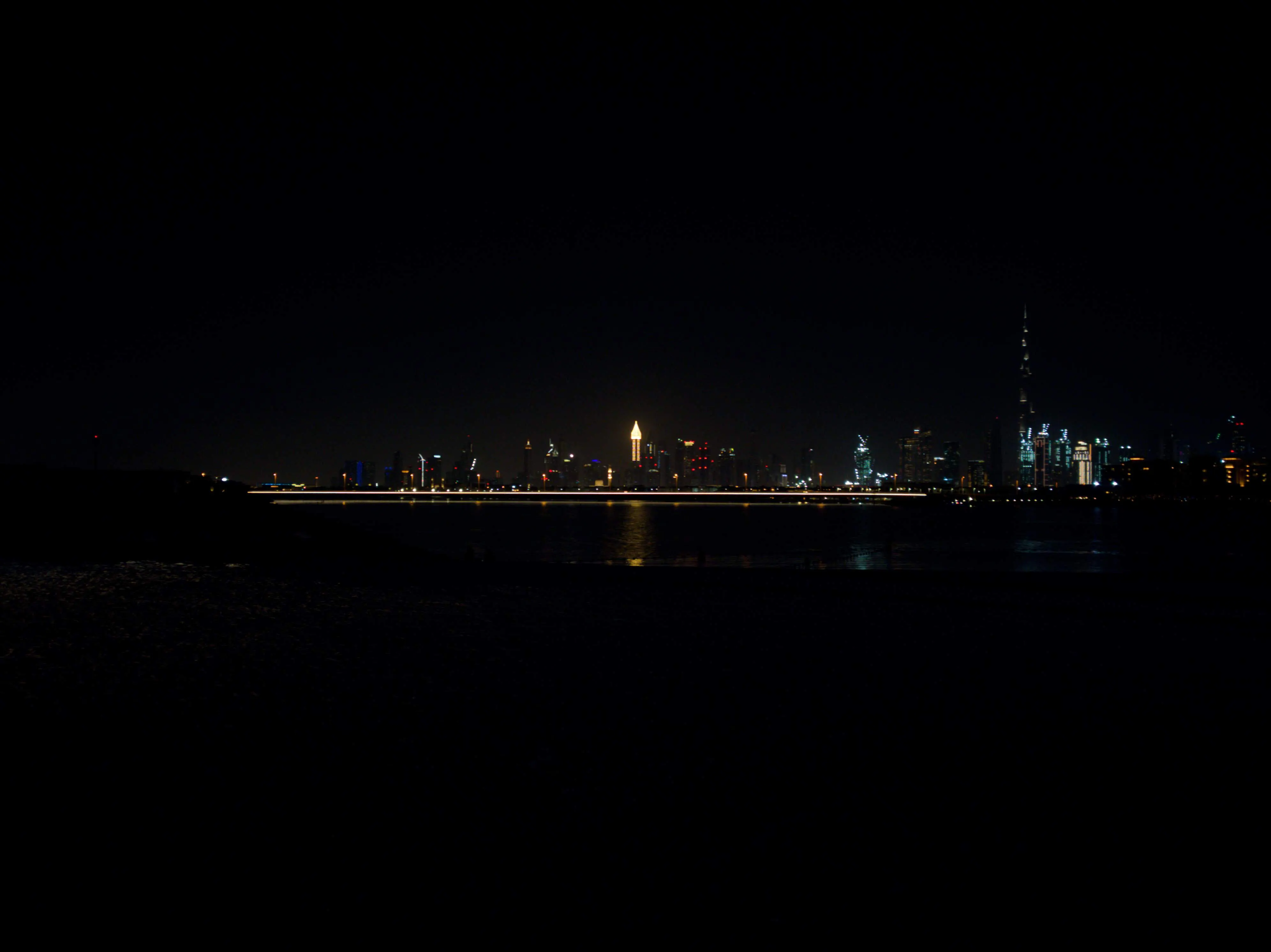 A long exposure of the Dubai skyline at night seen across dark water, the Burj Khalifa rising on the right and a streak of road lights cutting through the middle.