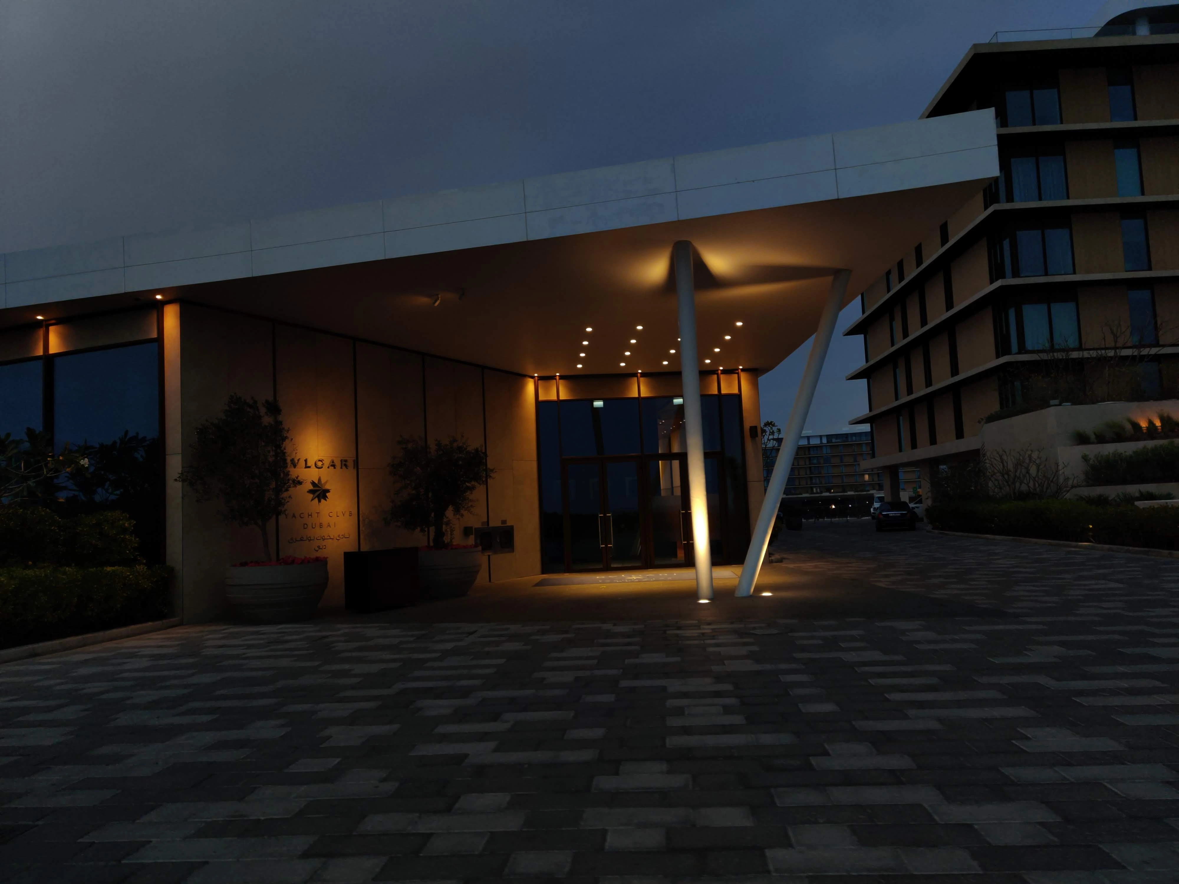 A low-lit modernist building entrance at dusk, warm interior lighting spilling onto a stone-paved drive under a deep blue sky.