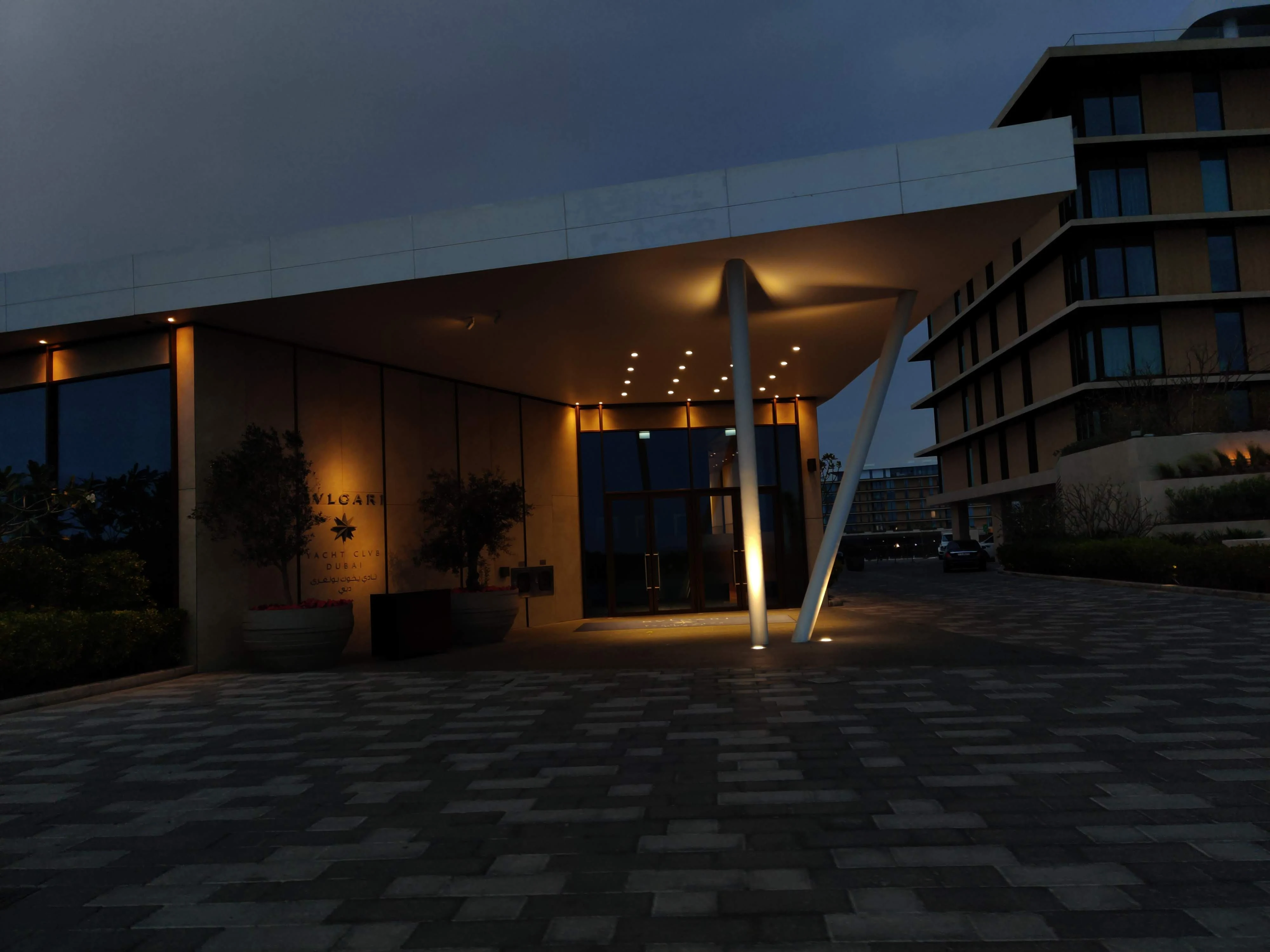 A low-lit modernist building entrance at dusk, warm interior lighting spilling onto a stone-paved drive under a deep blue sky.