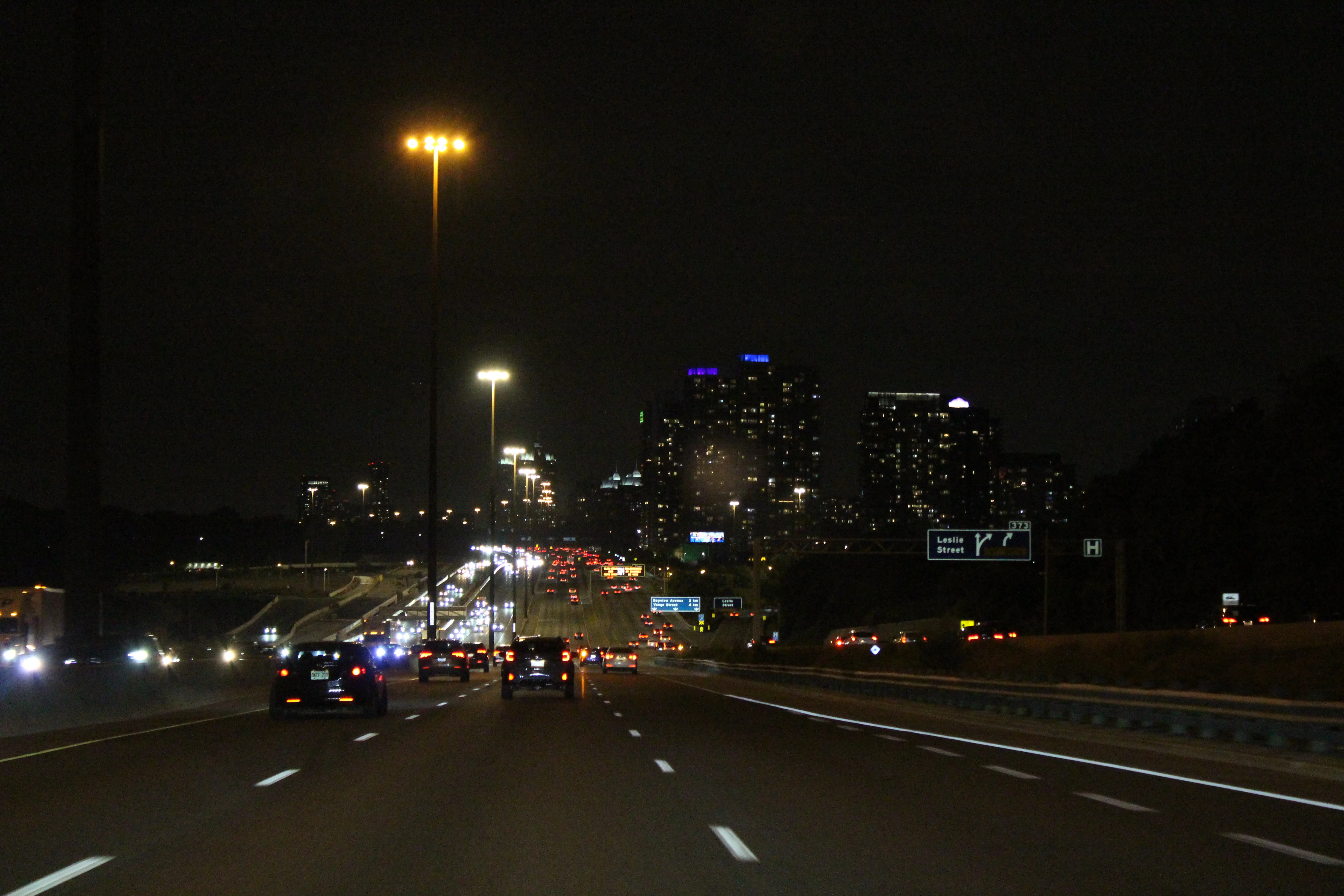 A night highway running toward a cluster of lit towers, tail lights strung out across the lanes ahead and a green overhead sign for Leslie Street.