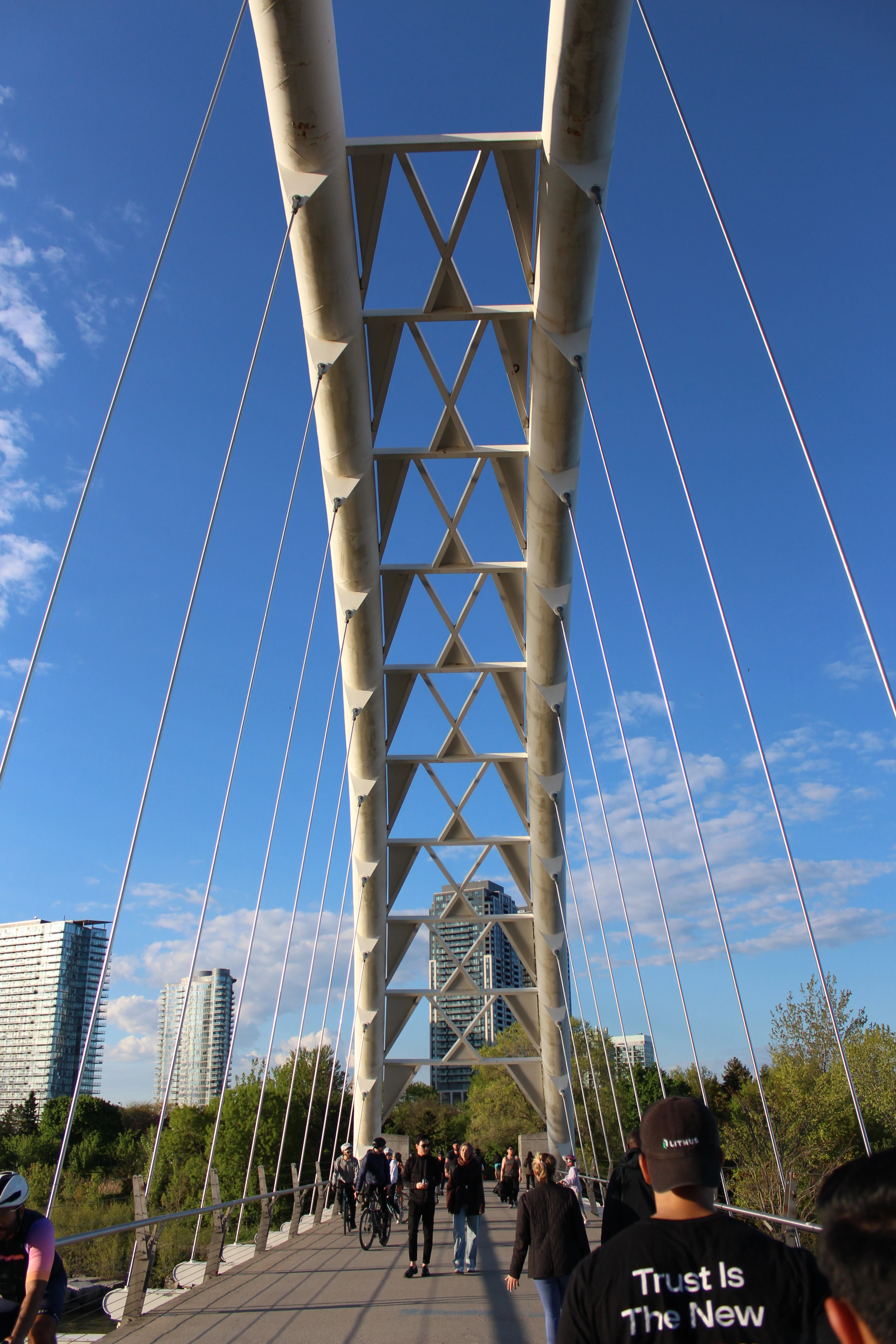 Looking up the steel truss of a cable-stayed pedestrian bridge, with the city behind it.