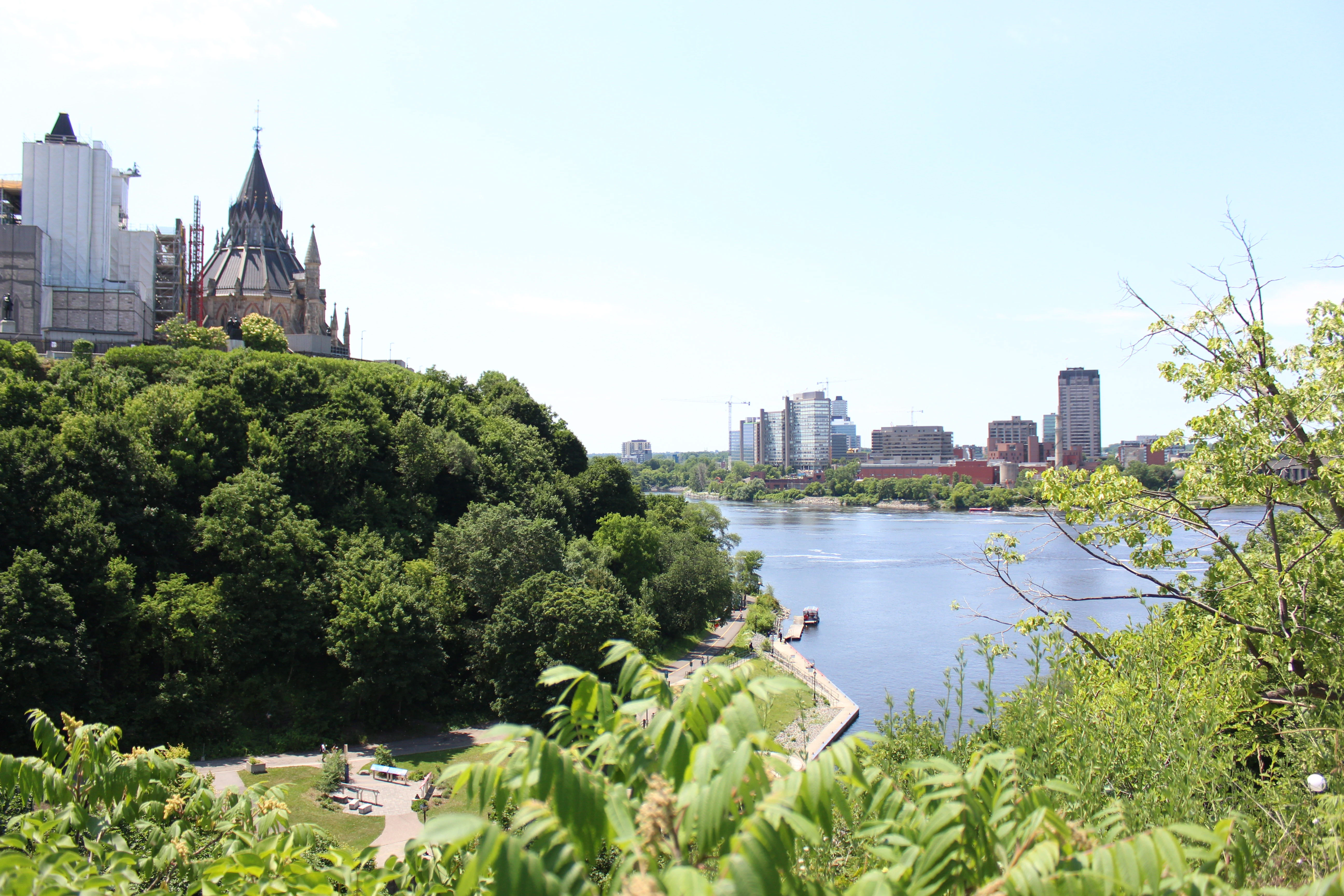 View from a hilltop showing the green copper roof of the Library of Parliament on the left, with the Ottawa River curving below and the Gatineau skyline beyond.
