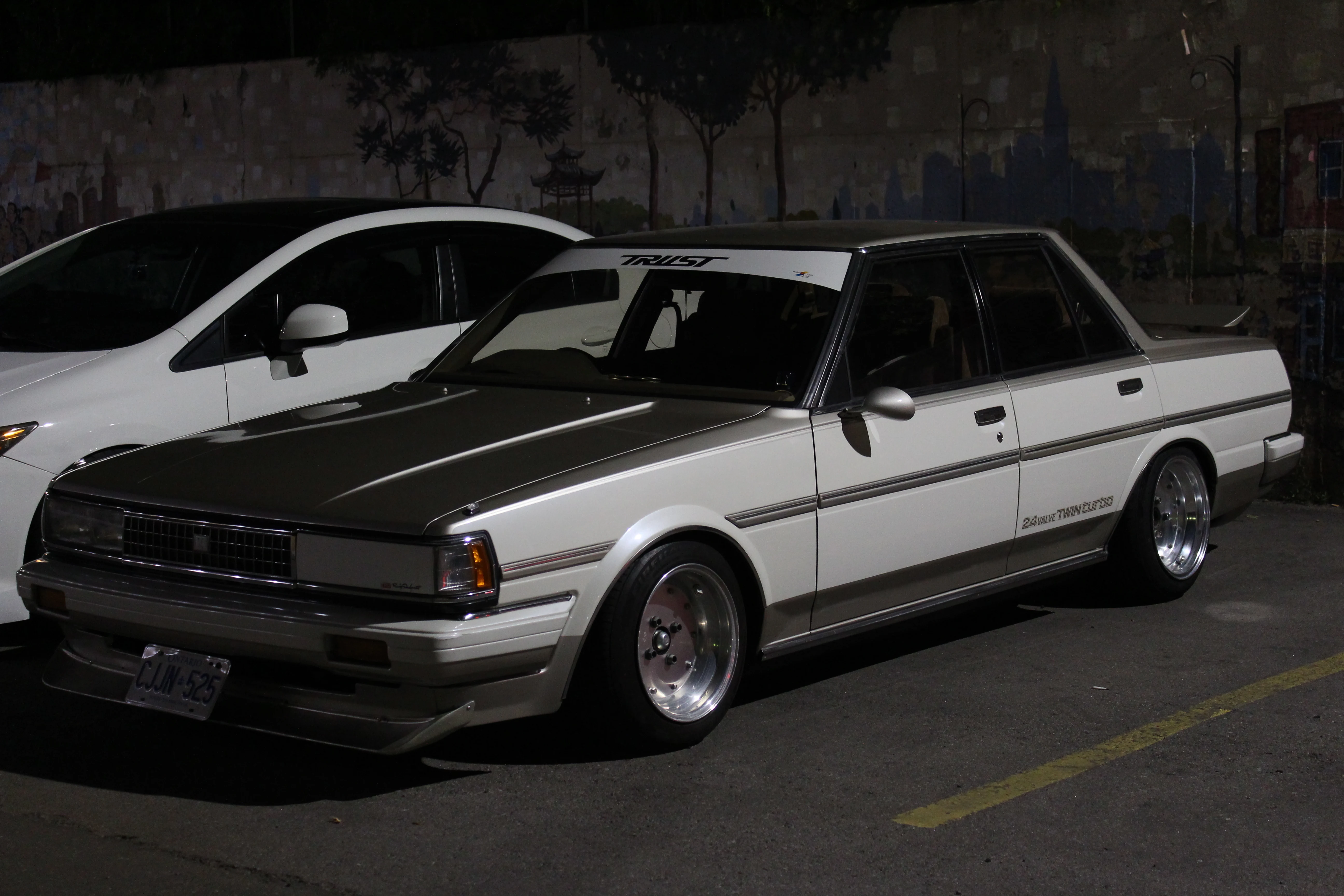 A lowered white and grey Japanese sedan parked at night against a painted mural wall, polished wheels catching the streetlight.