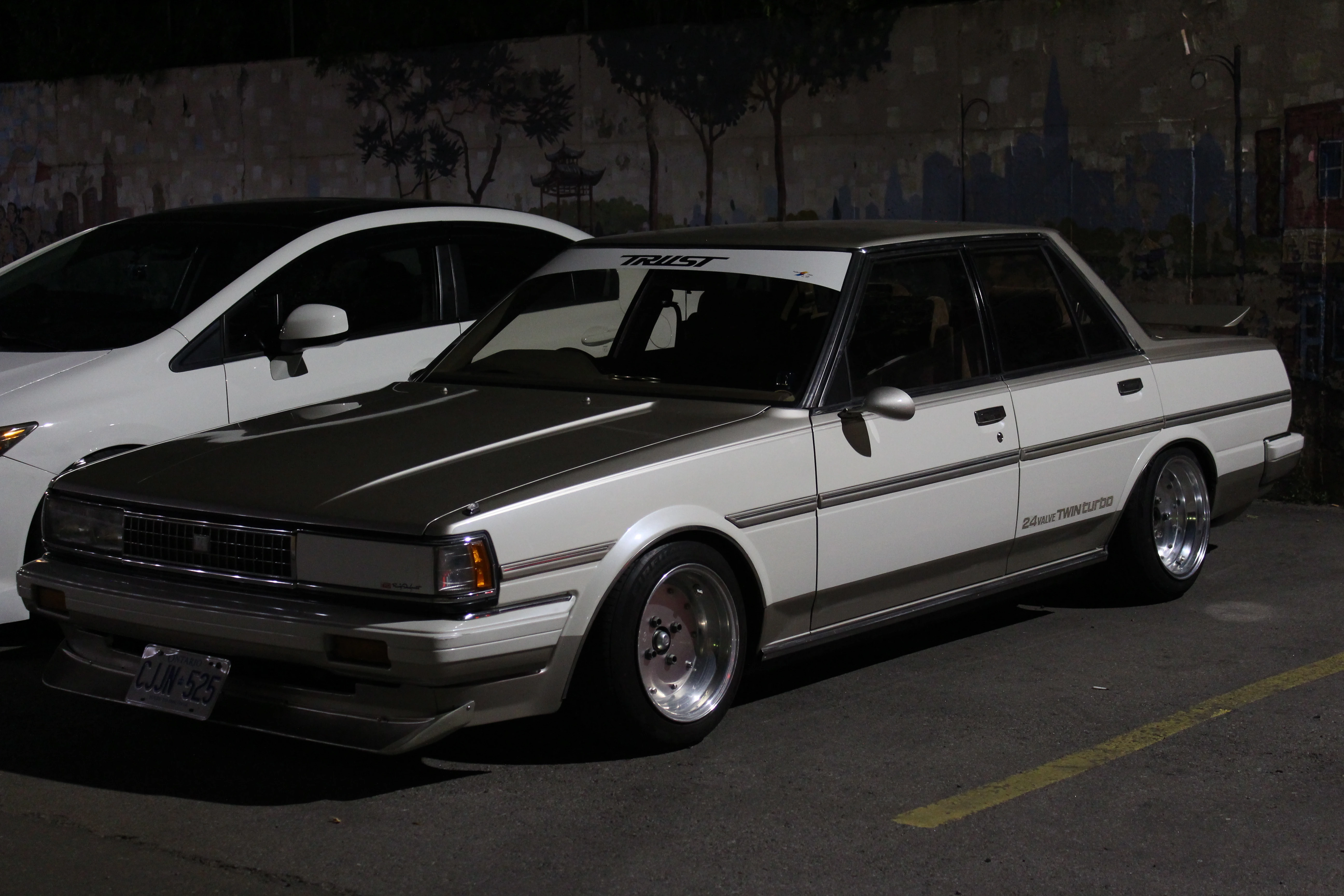 A lowered white and grey Japanese sedan parked at night against a painted mural wall, polished wheels catching the streetlight.