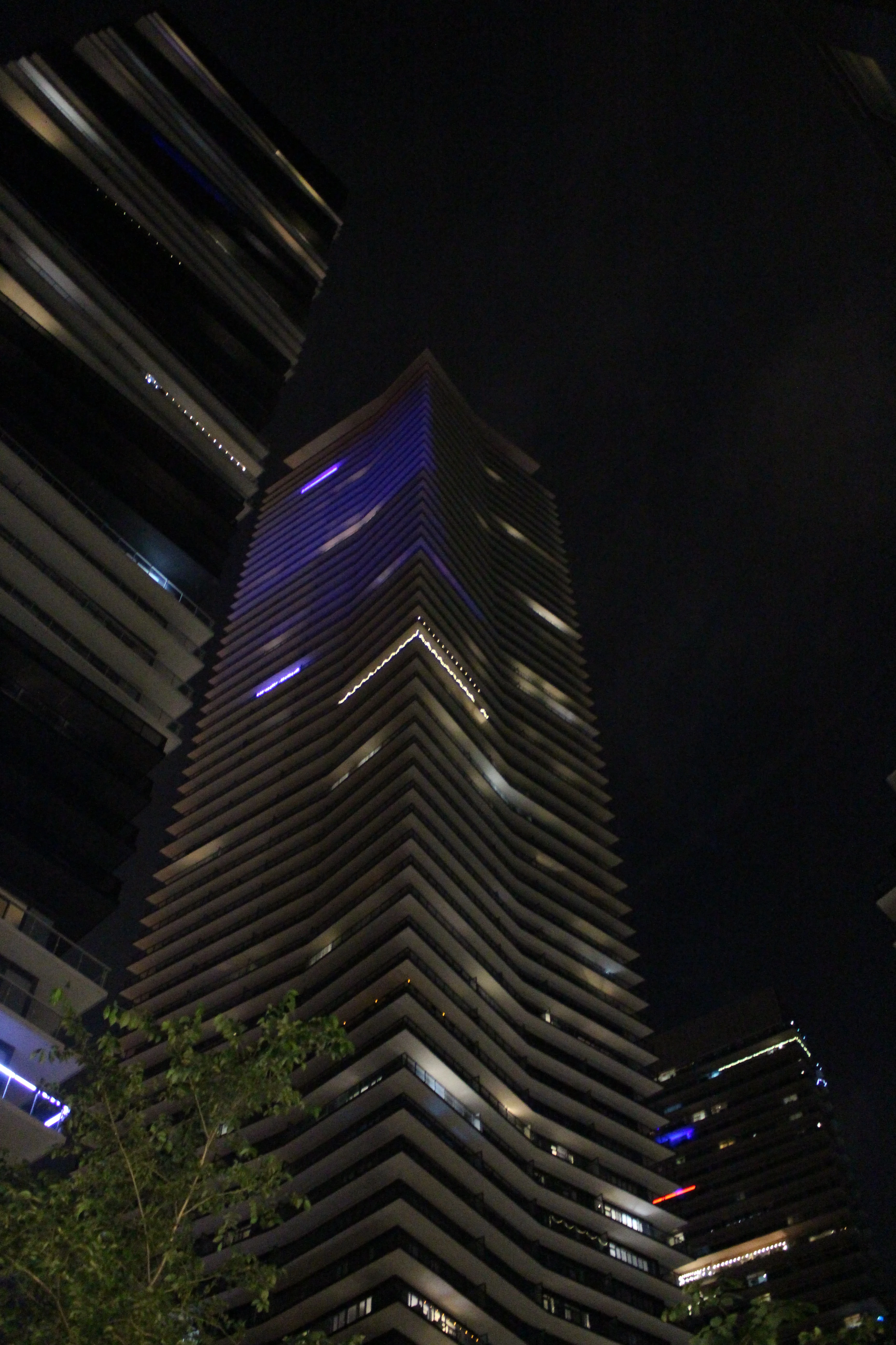 A tall tower seen from below at night, ribbed facade fanning out toward a faint blue glow at the crown.