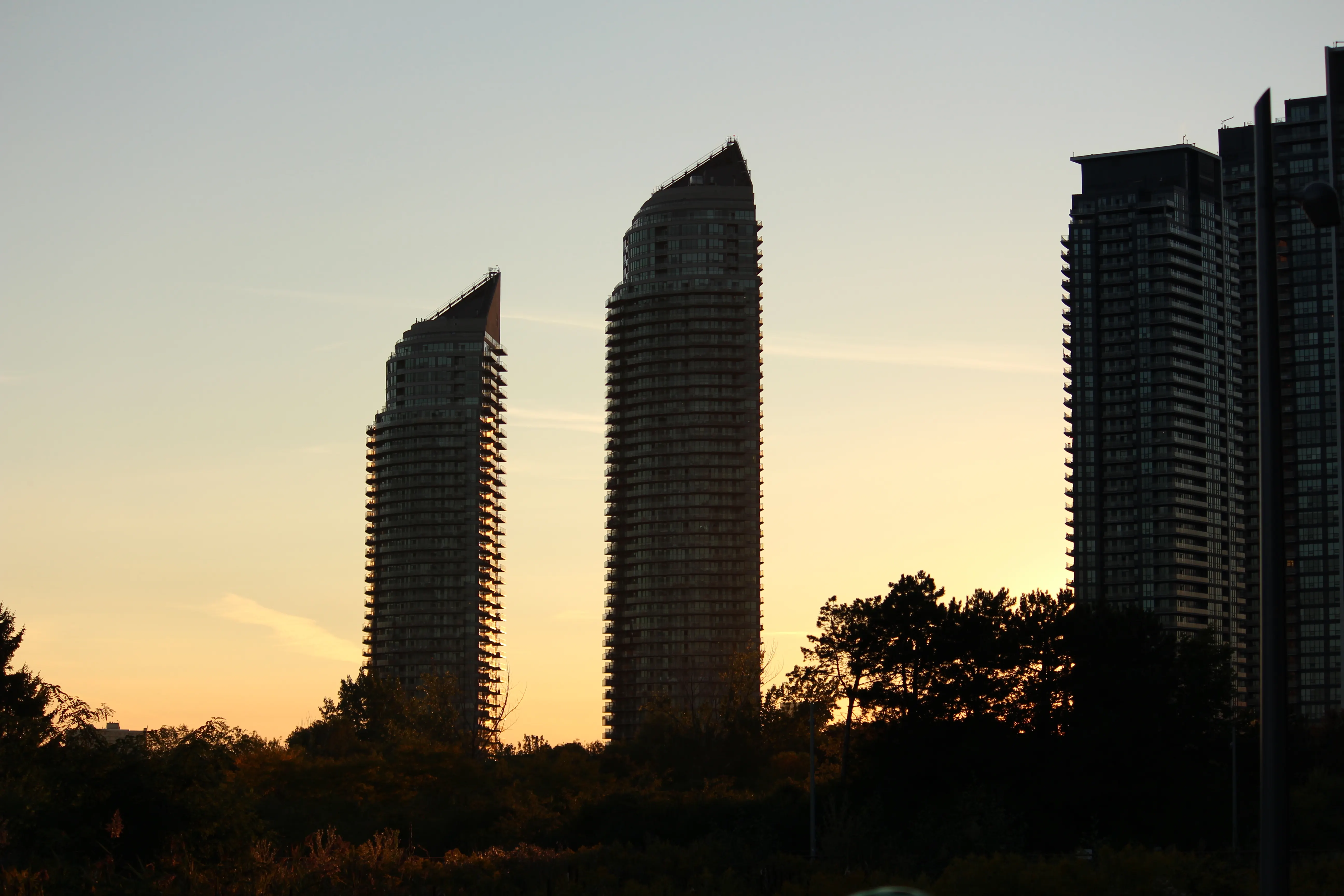Two curved condominium towers silhouetted against a warm sunset sky, tree line low across the foreground.