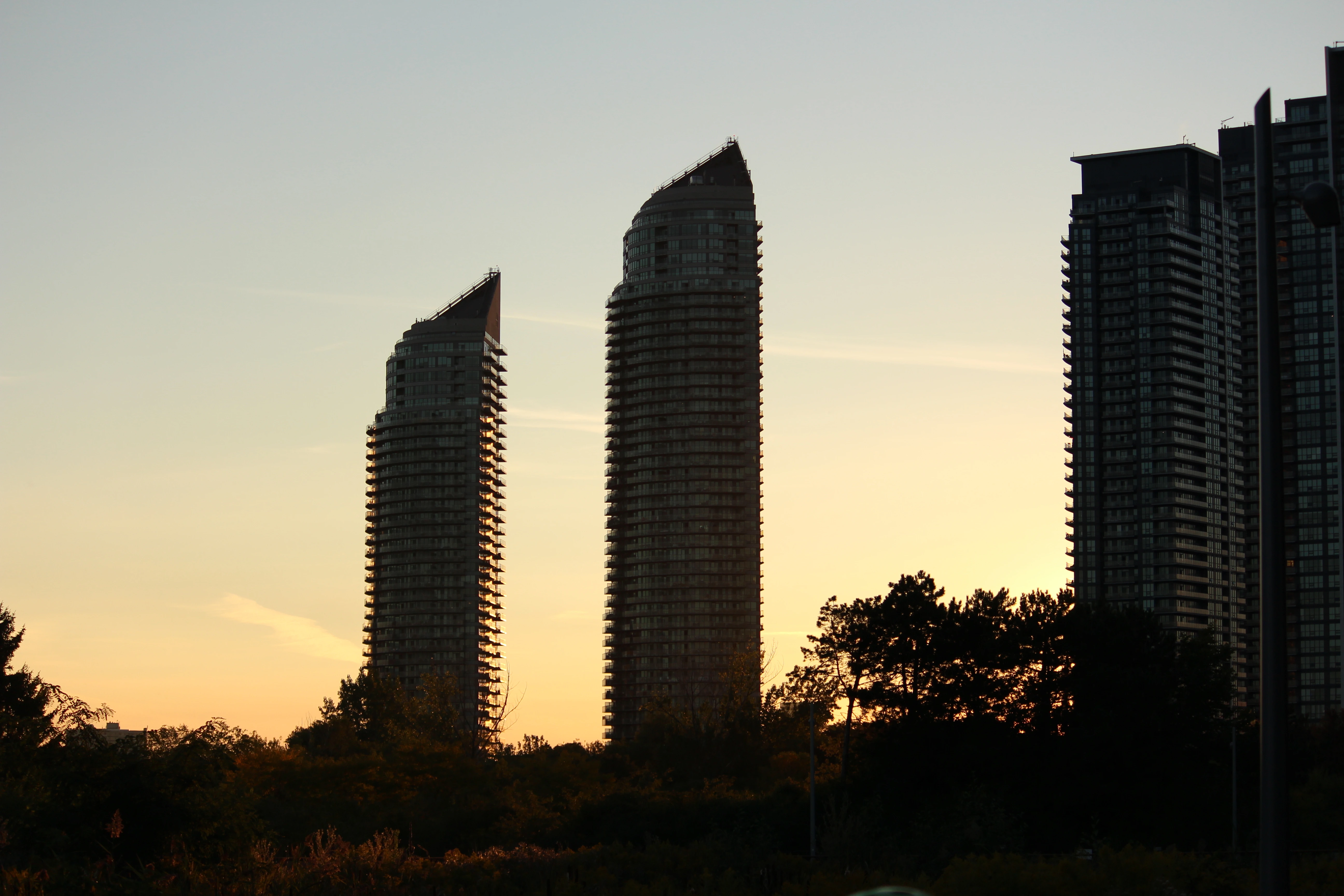 Two curved condominium towers silhouetted against a warm sunset sky, tree line low across the foreground.