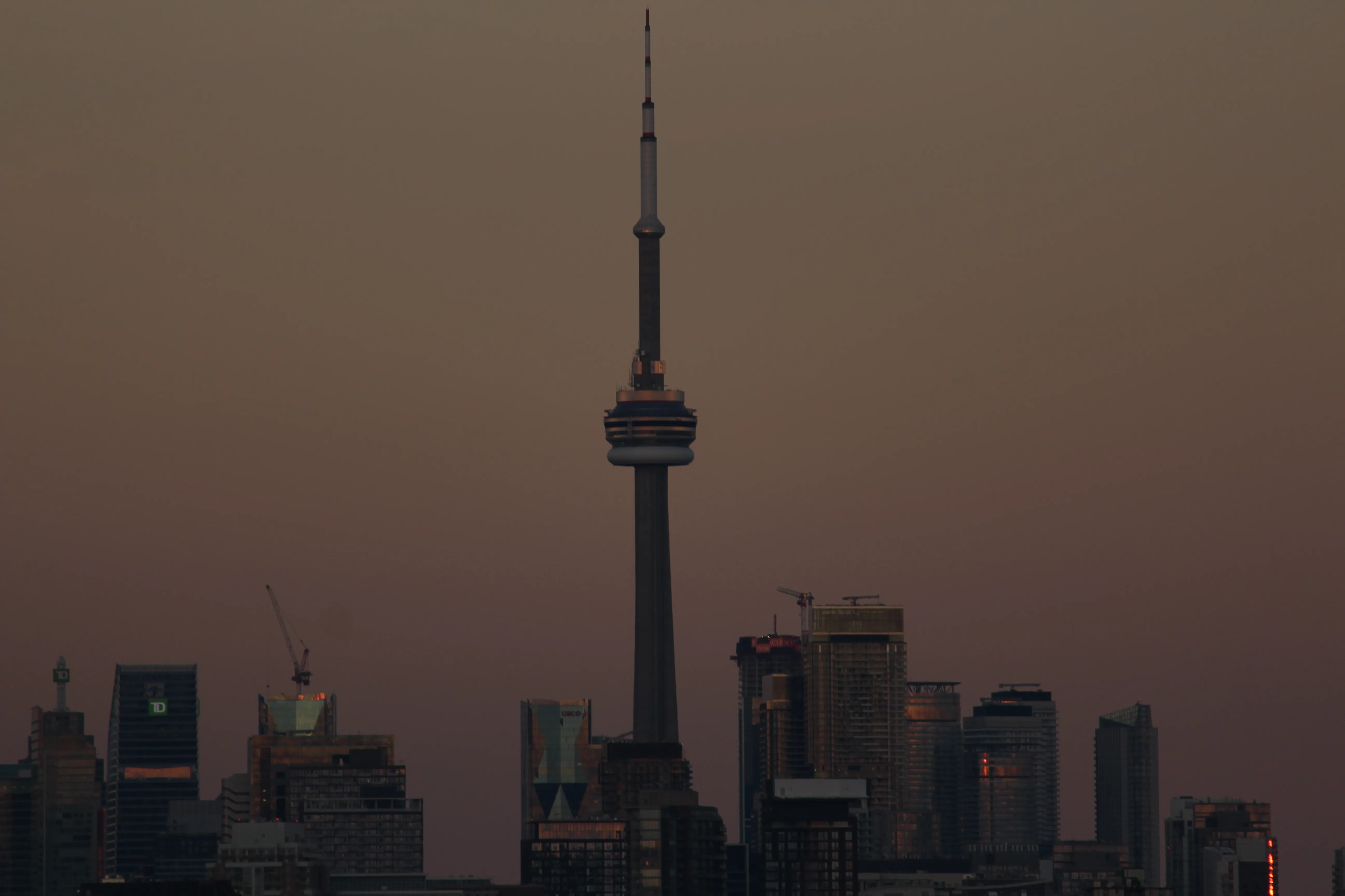 The CN Tower silhouetted against a hazy amber-pink dusk sky, the Toronto skyline below catching the last light.