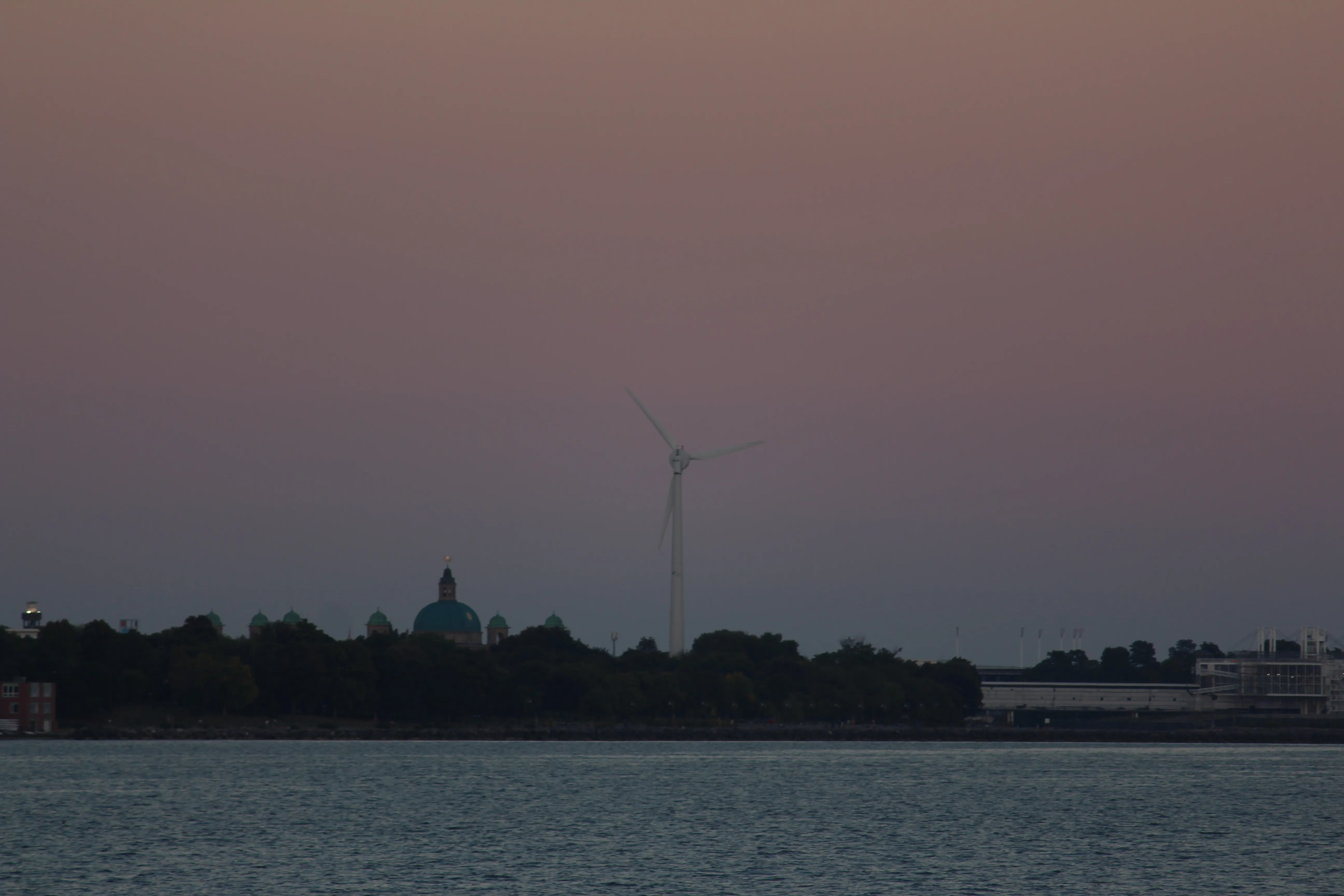 A wind turbine and a green church dome silhouetted against a dusty pink dusk sky over water.