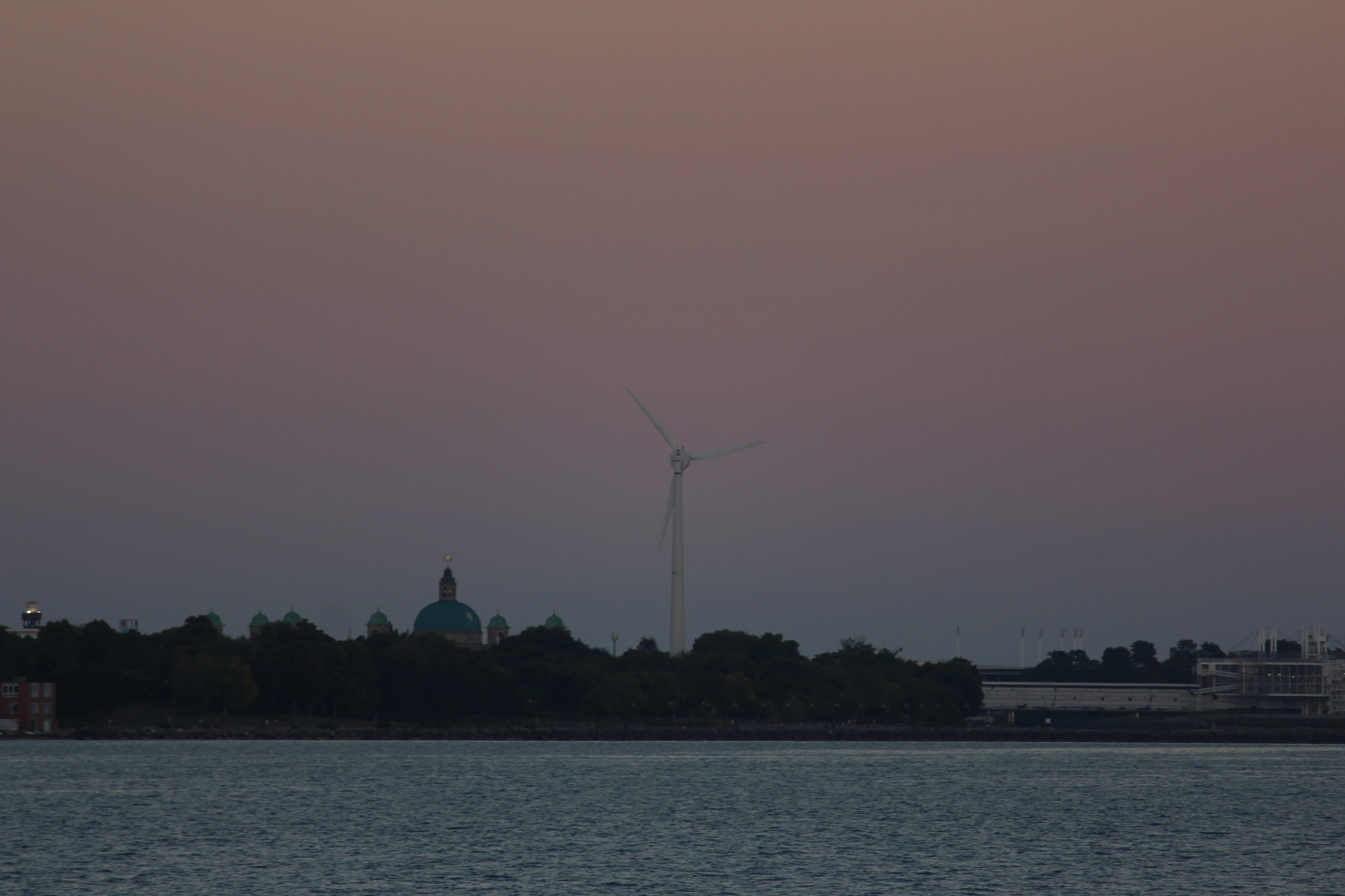 A wind turbine and a green church dome silhouetted against a dusty pink dusk sky over water.