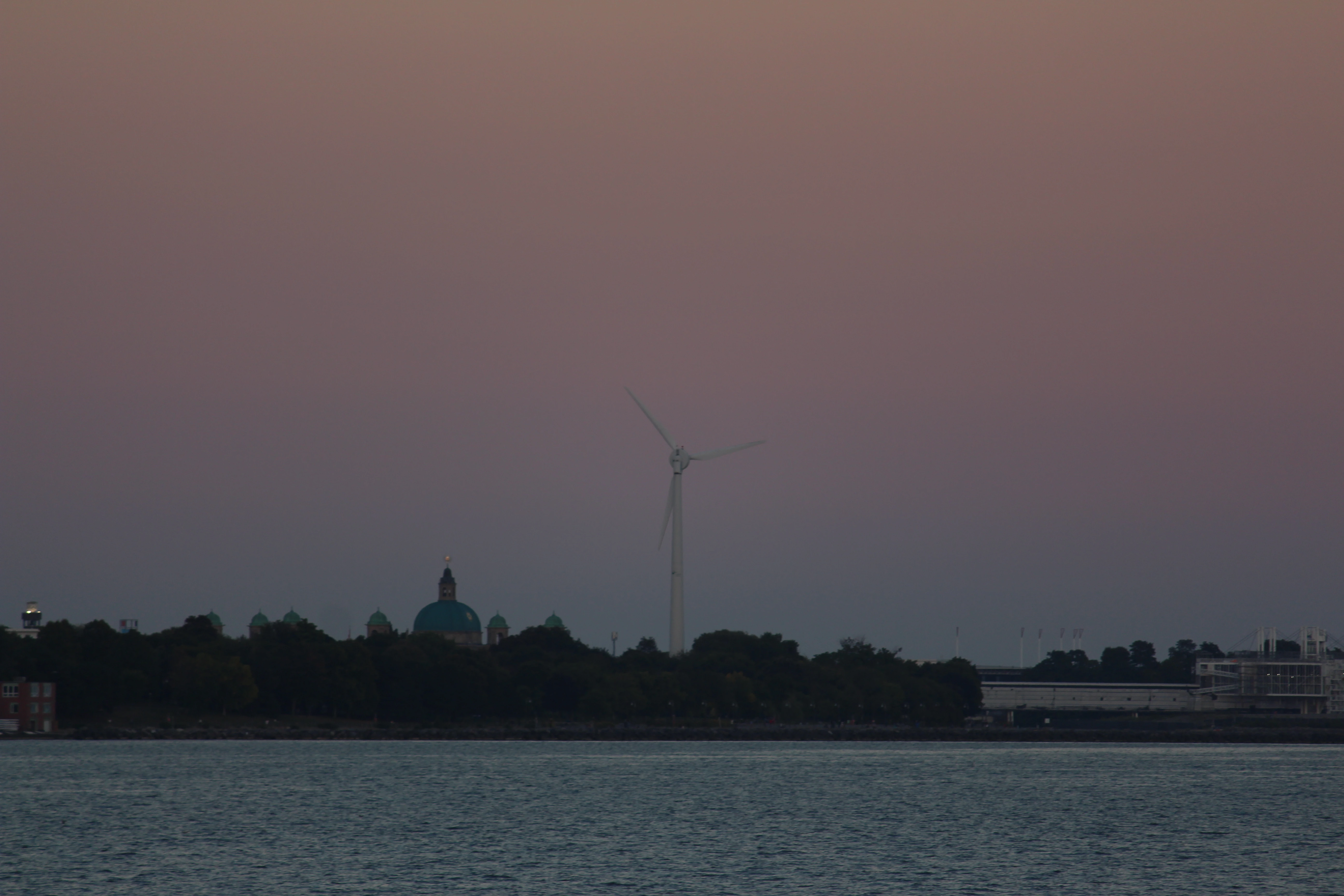 A wind turbine and a green church dome silhouetted against a dusty pink dusk sky over water.