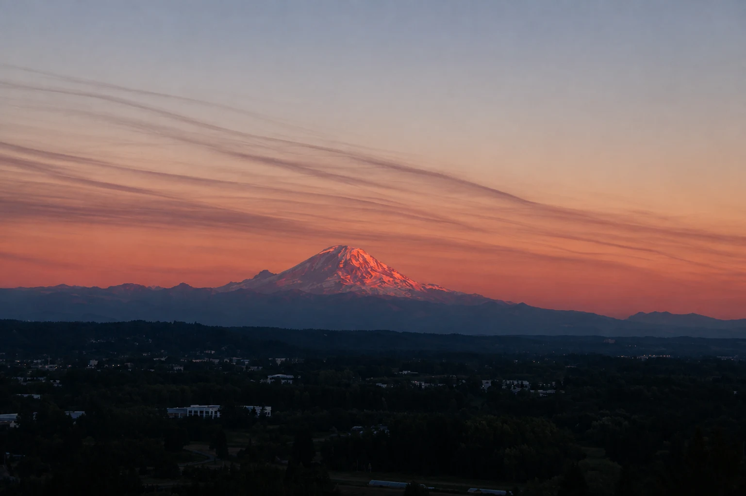 Mount Rainier catching pink sunset light above a darkening, forested valley under a streaked orange and violet sky.