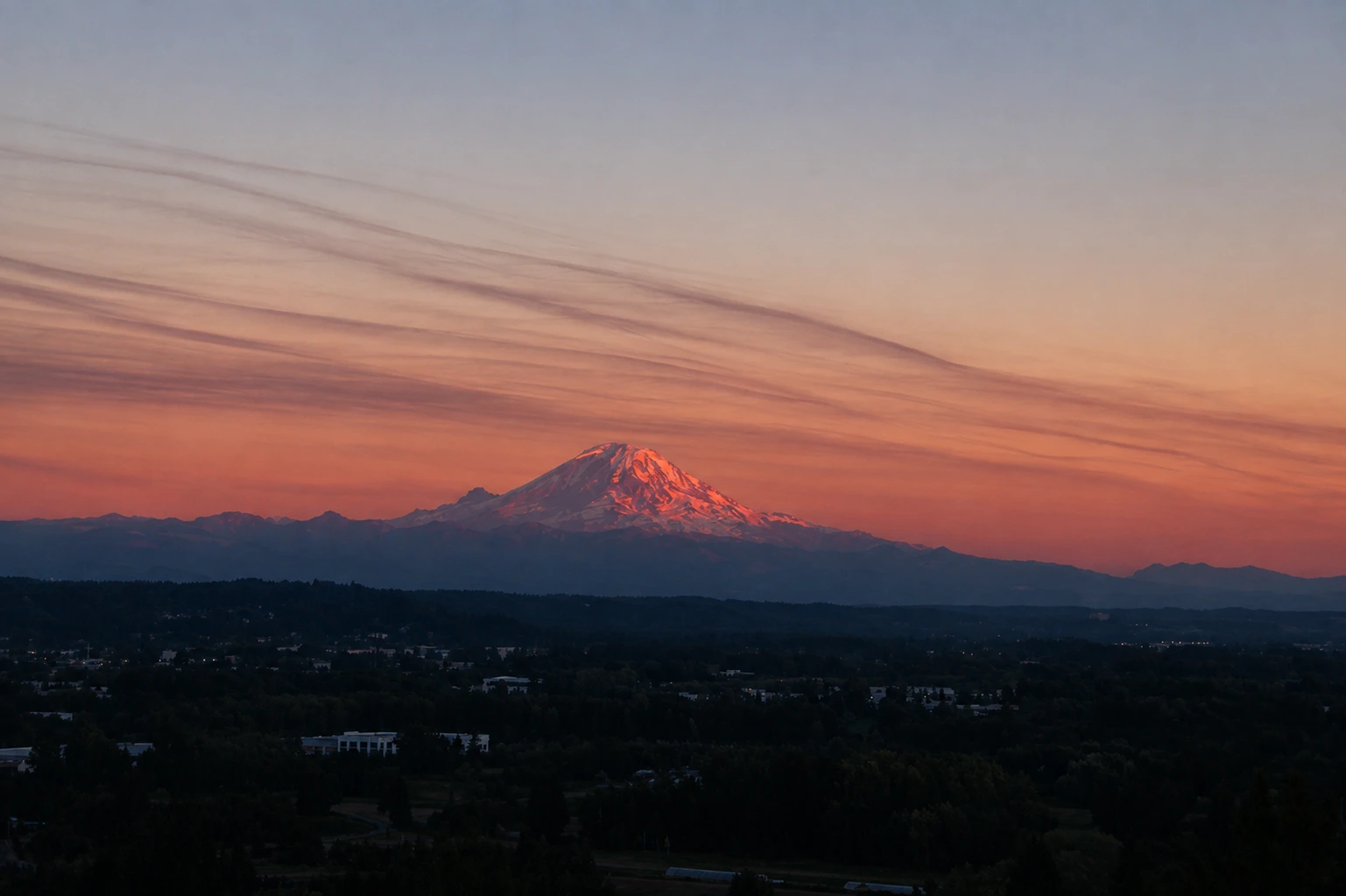Mount Rainier catching pink sunset light above a darkening, forested valley under a streaked orange and violet sky.