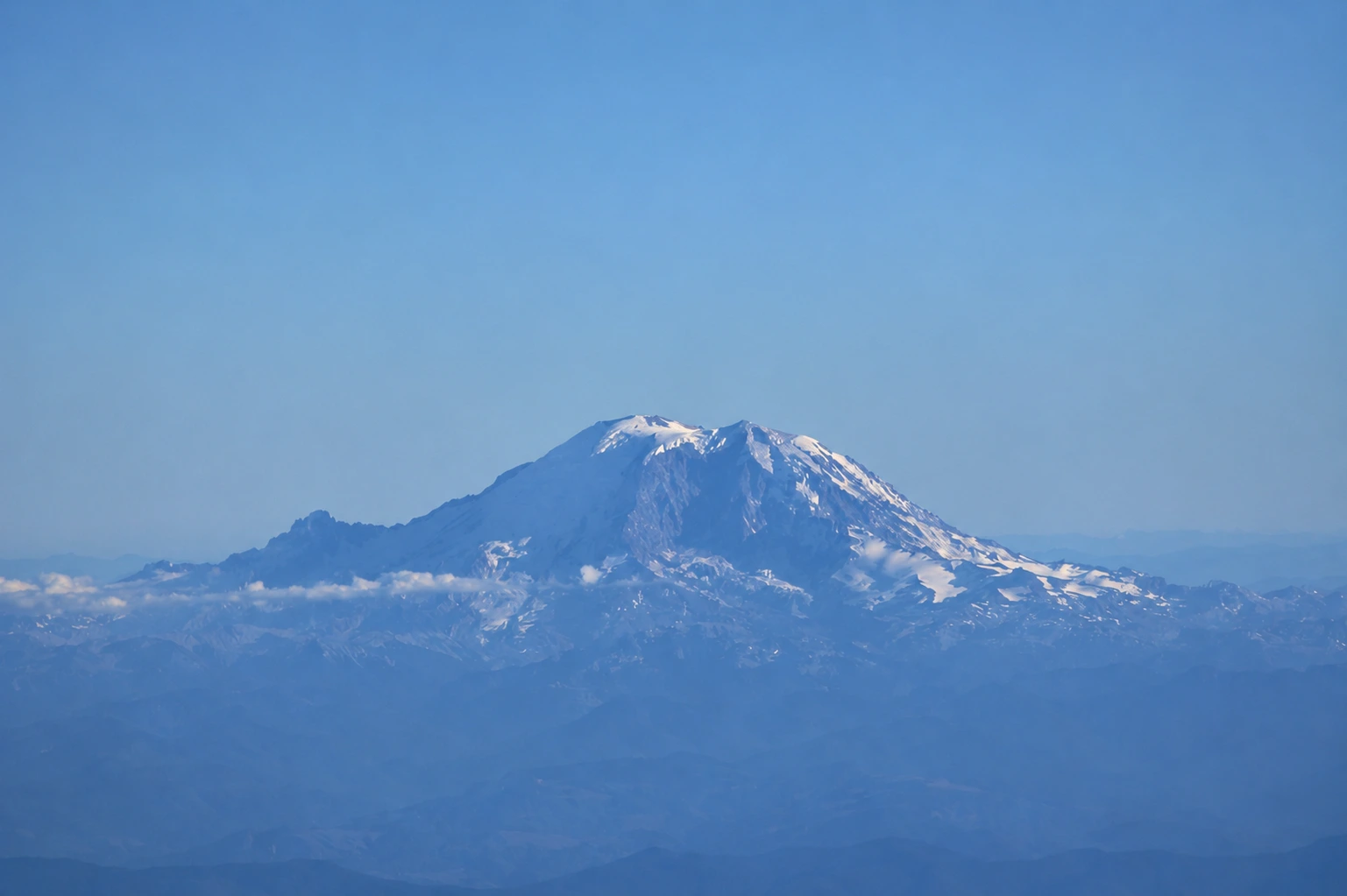 Mount Rainier's snow-capped summit rising above layered foothills and haze under a clear sky, seen from the air.