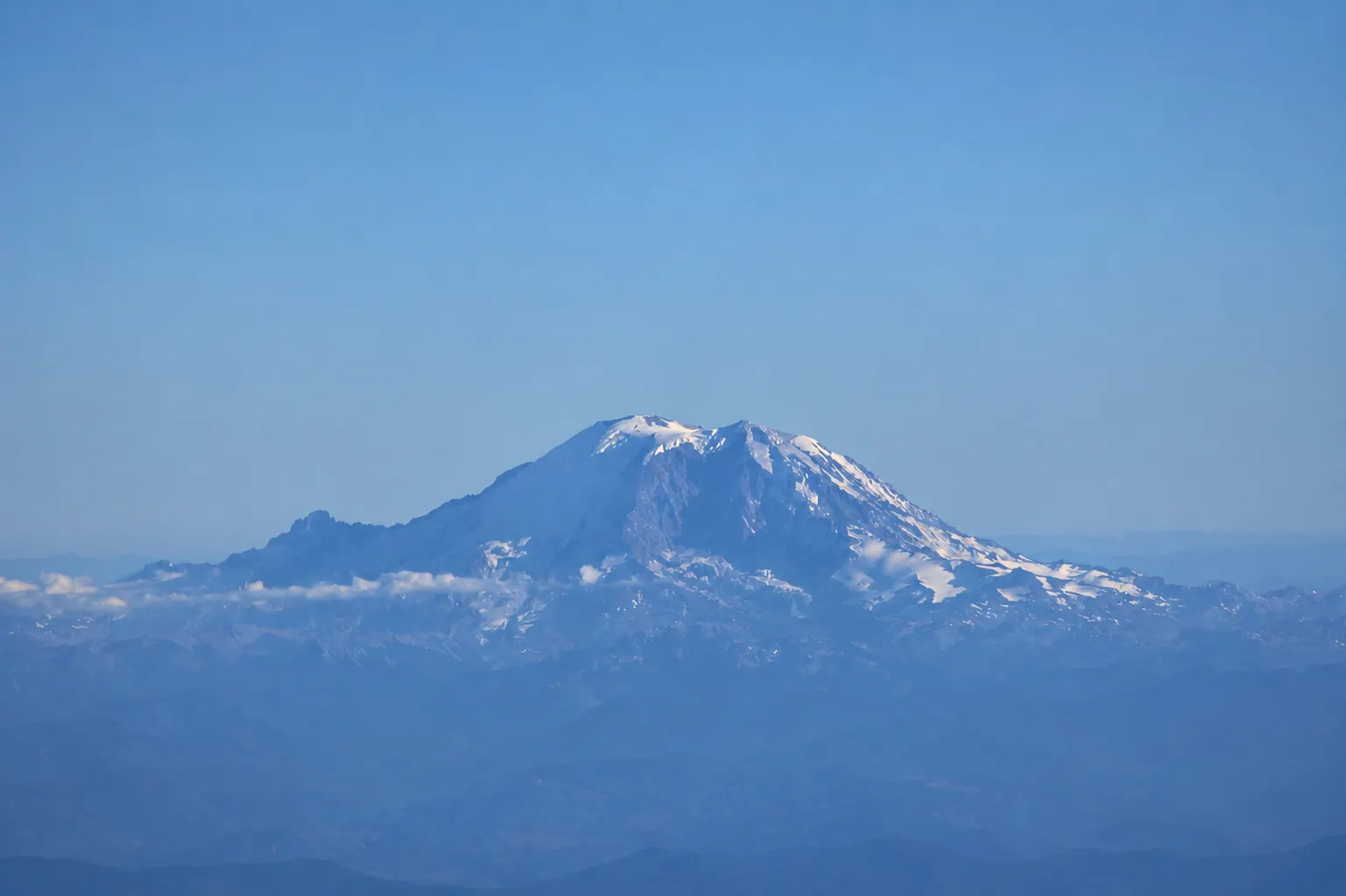 Mount Rainier's snow-capped summit rising above layered foothills and haze under a clear sky, seen from the air.