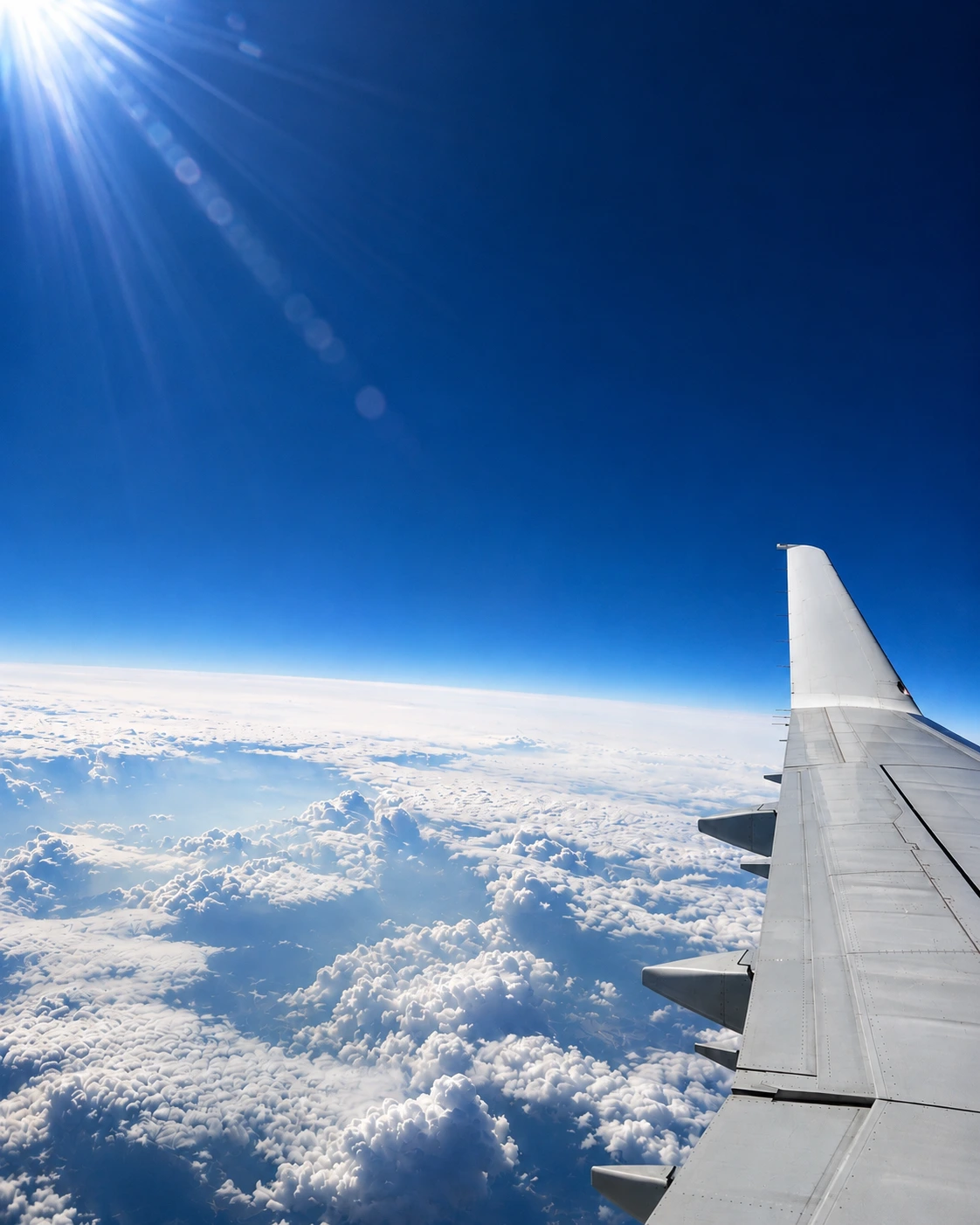 An airliner's wing reaching into deep blue sky, the sun flaring at the top of the frame above a wide floor of cloud.